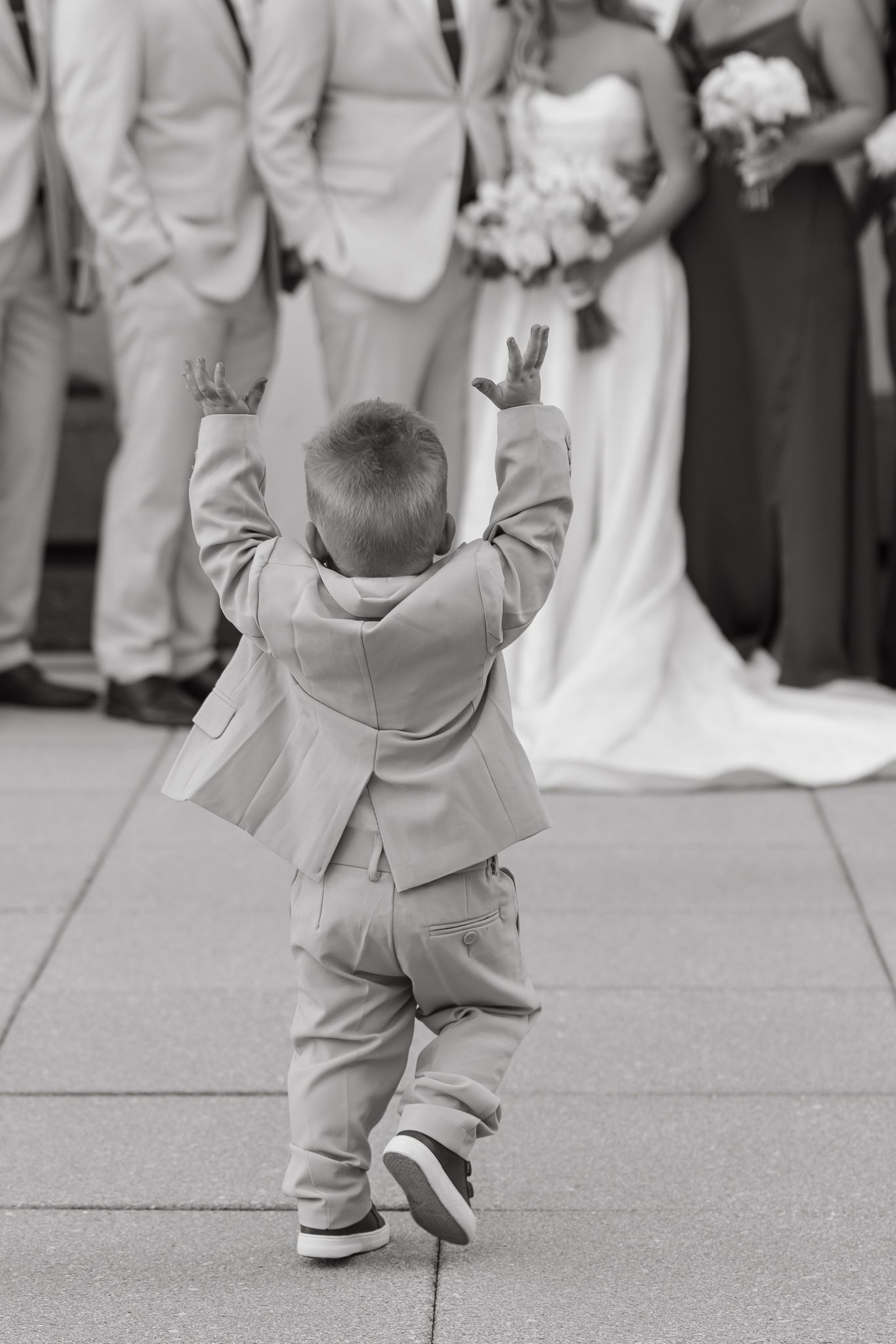 A black and white photo shows a small child in formal attire walking across a floor at a wedding ceremony.