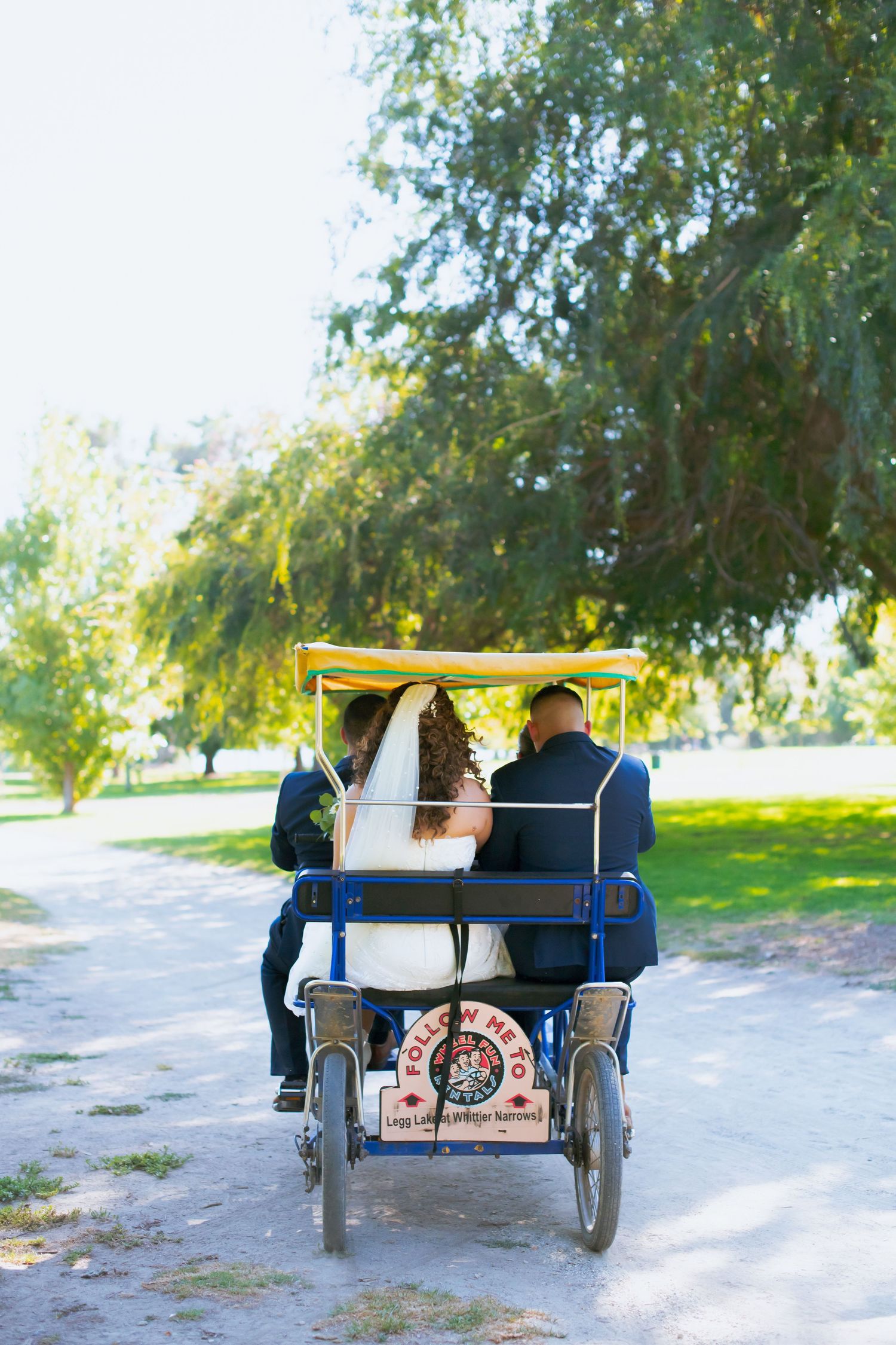 Wedding couple rides in decorative carriage under trees at outdoor venue Whittier Narrows Recreation Area.