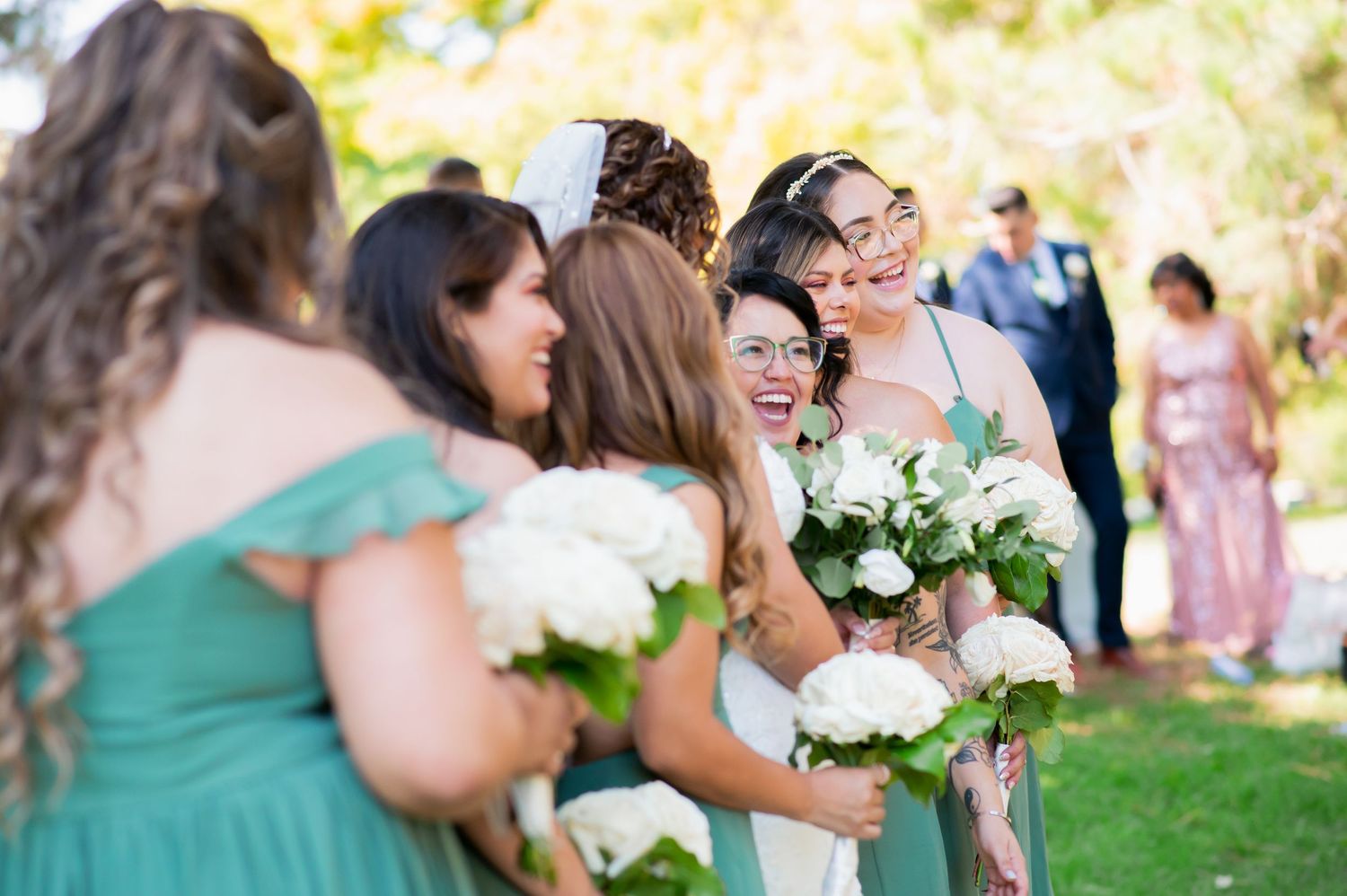 Wedding party laughs together while holding white floral bouquets outdoors.