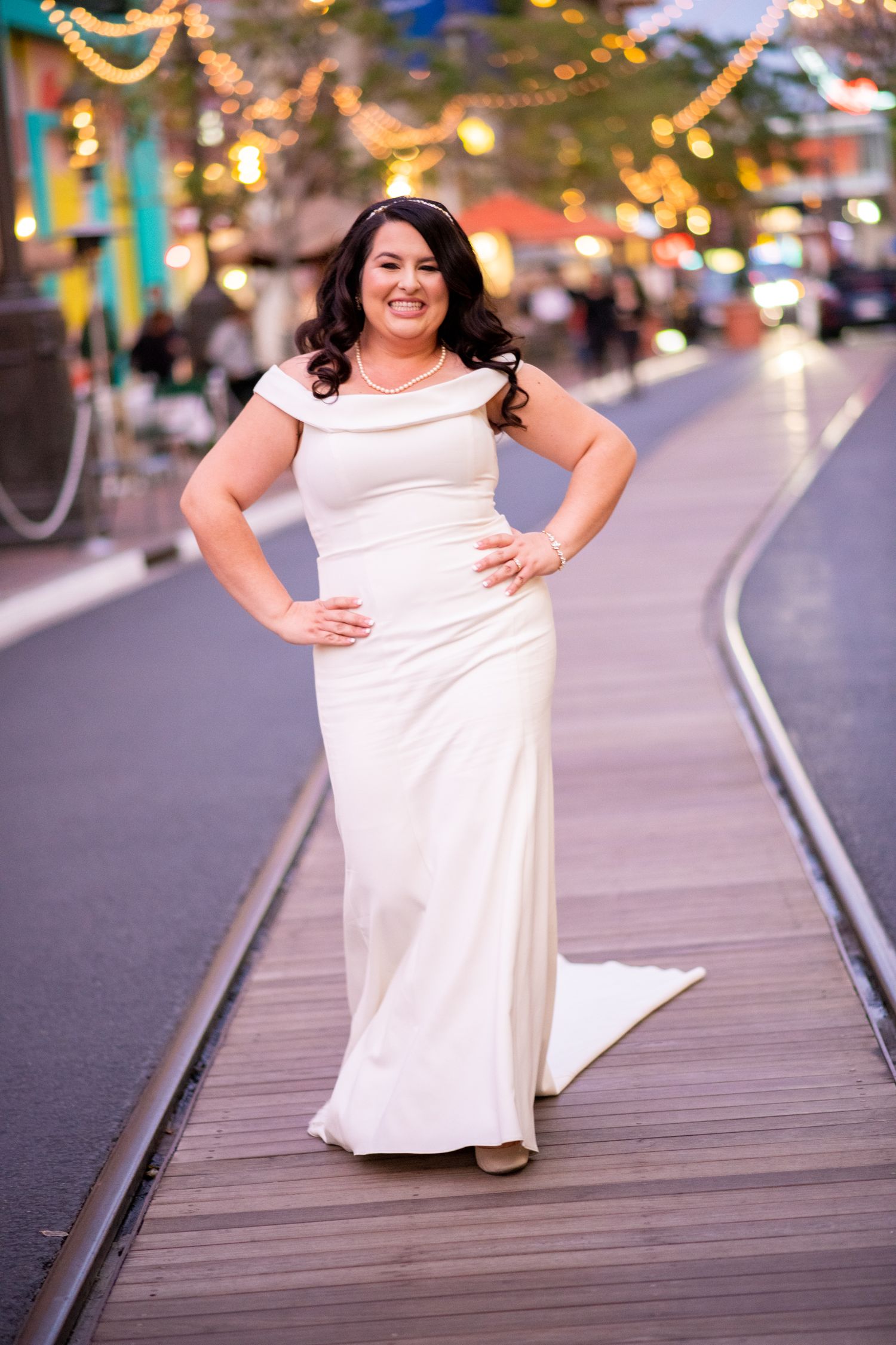 Bride in elegant white gown poses on boardwalk with twinkling lights in background.