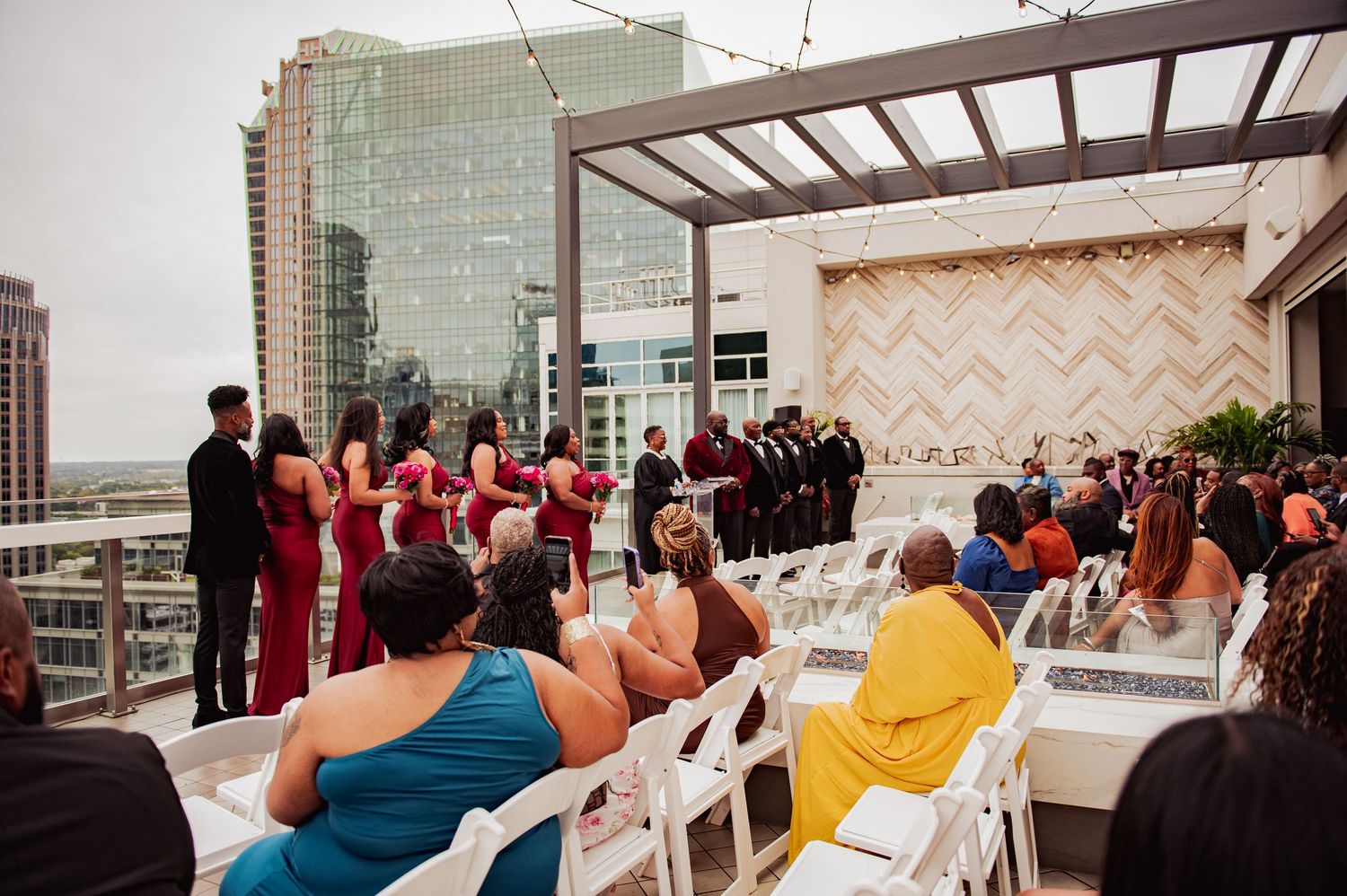 A wedding ceremony takes place on a rooftop venue with guests seated and a city skyline backdrop.