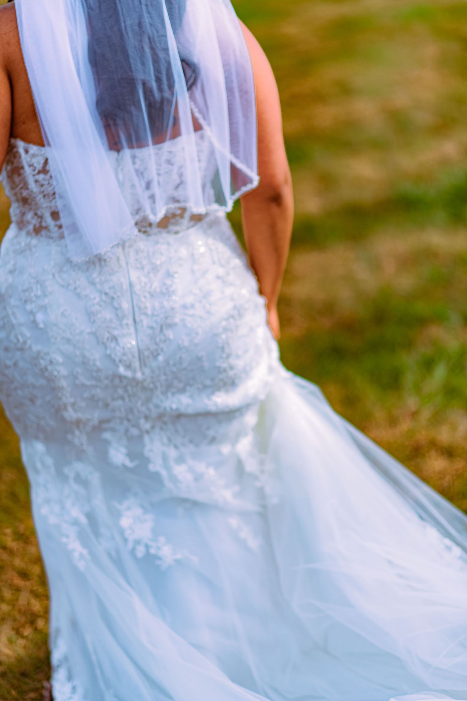 Close-up details of a white lace wedding dress with delicate embroidery and flowing tulle veil in an outdoor setting.