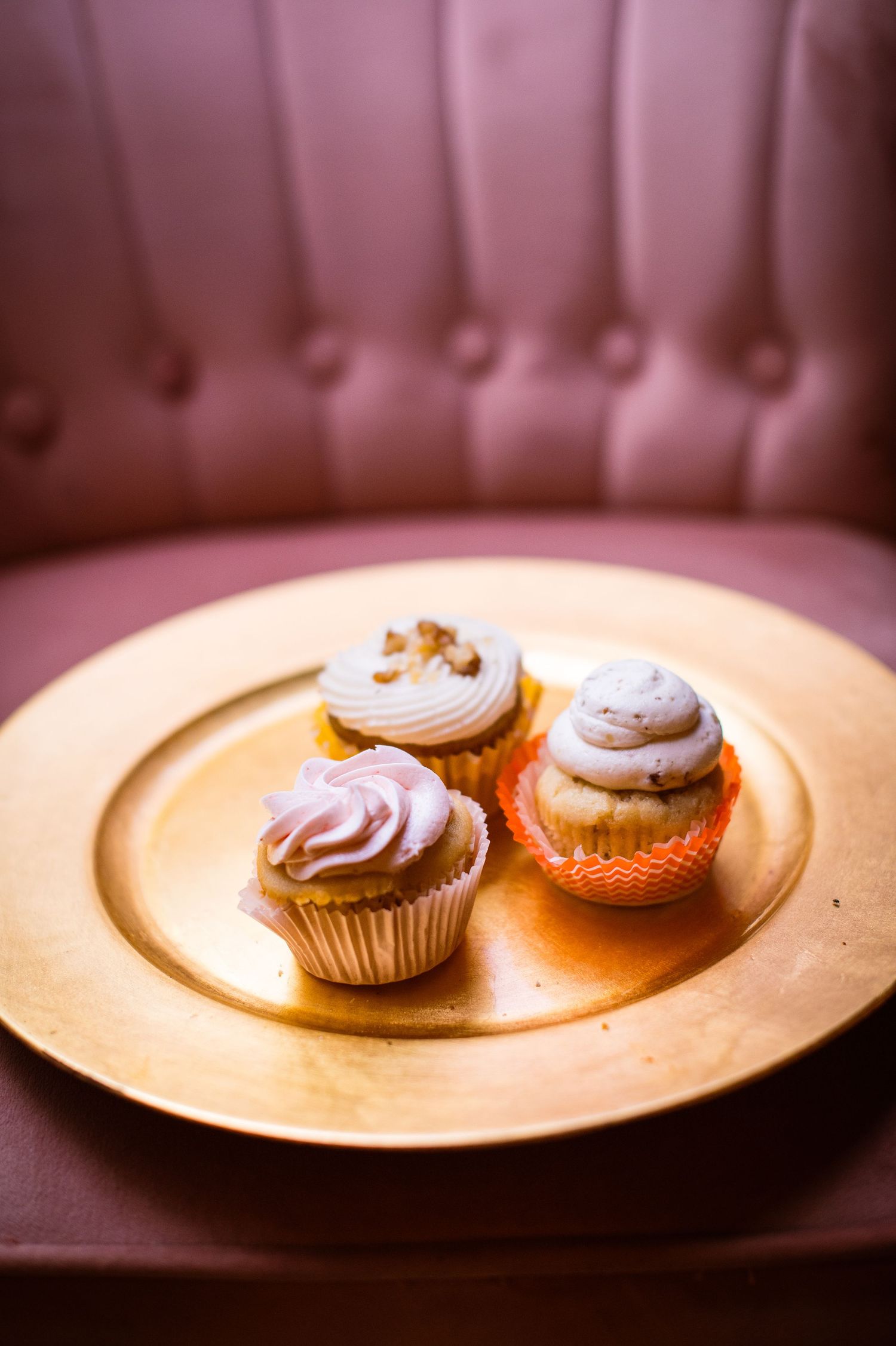 Three delicate cupcakes with swirled frosting displayed on a golden plate against pink upholstery.