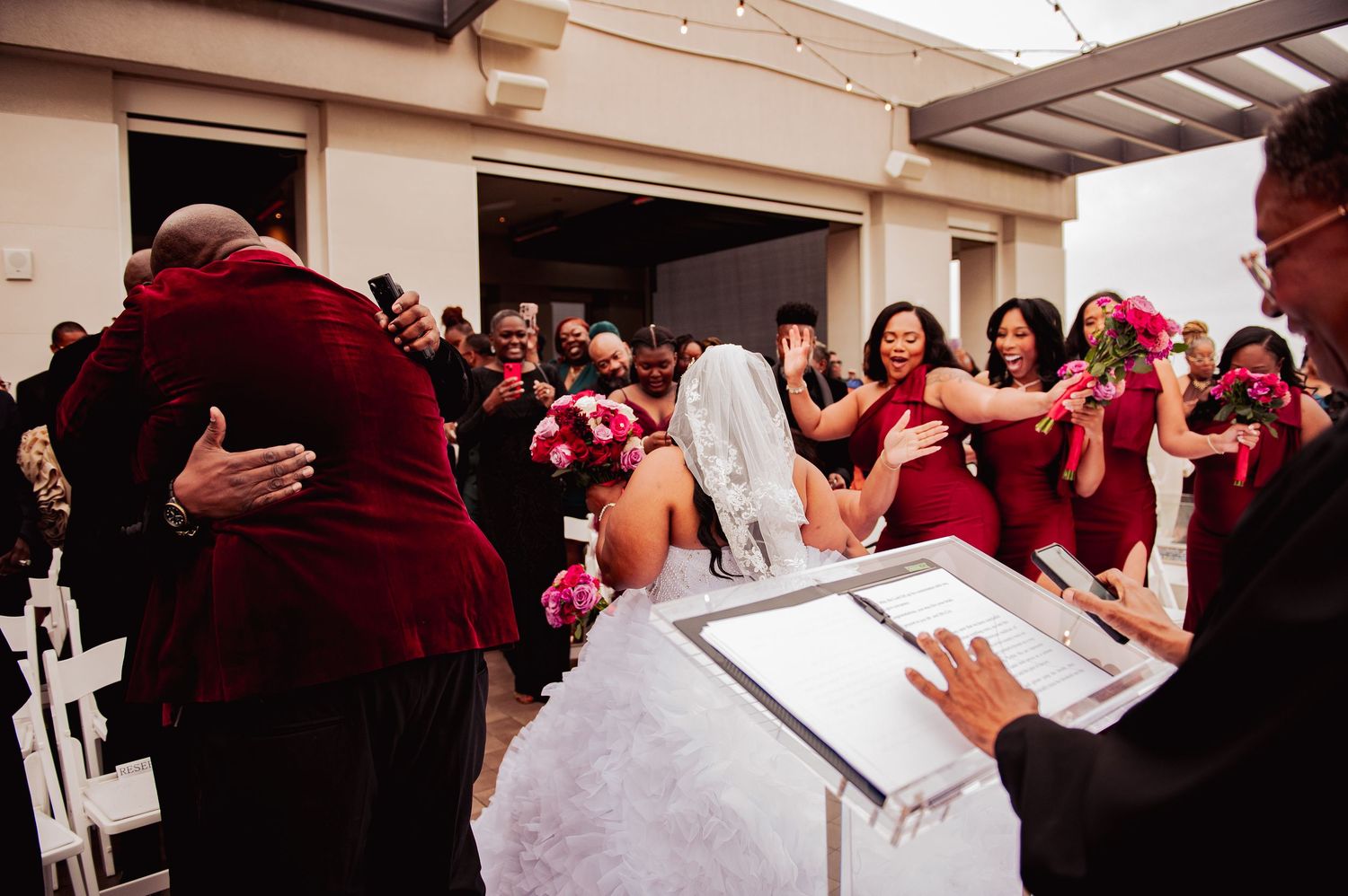 Wedding party in red dresses celebrates during ceremony signing with bride in white gown.