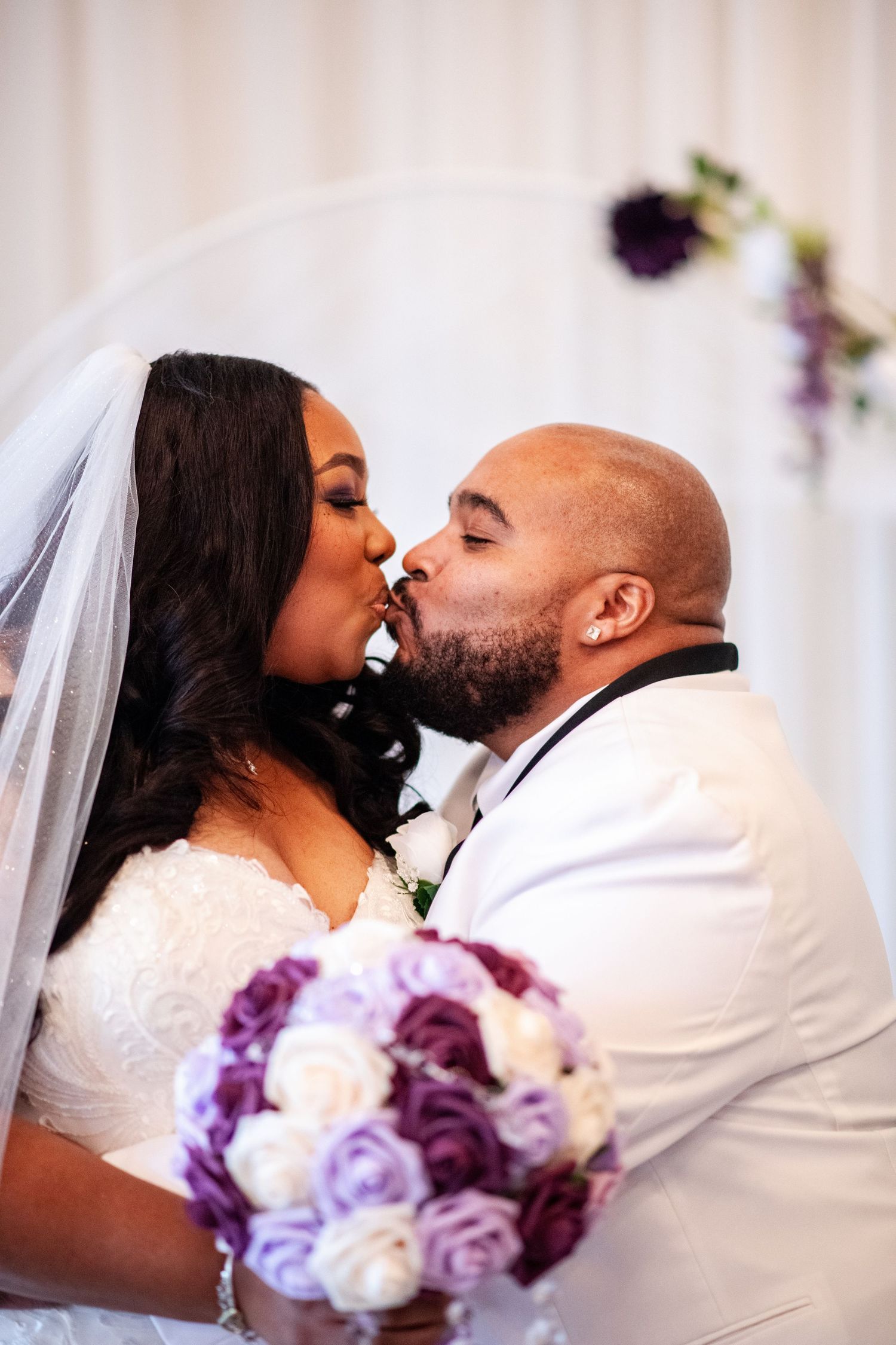Romantic wedding ceremony kiss moment with purple and white floral bouquet visible.