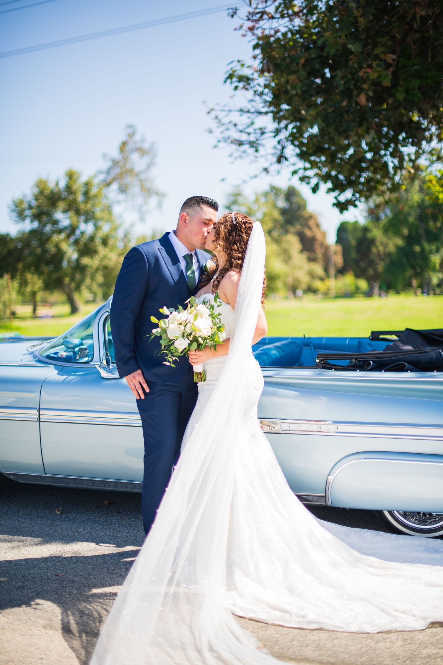 Romantic wedding portrait with couple against silver vintage car under trees.