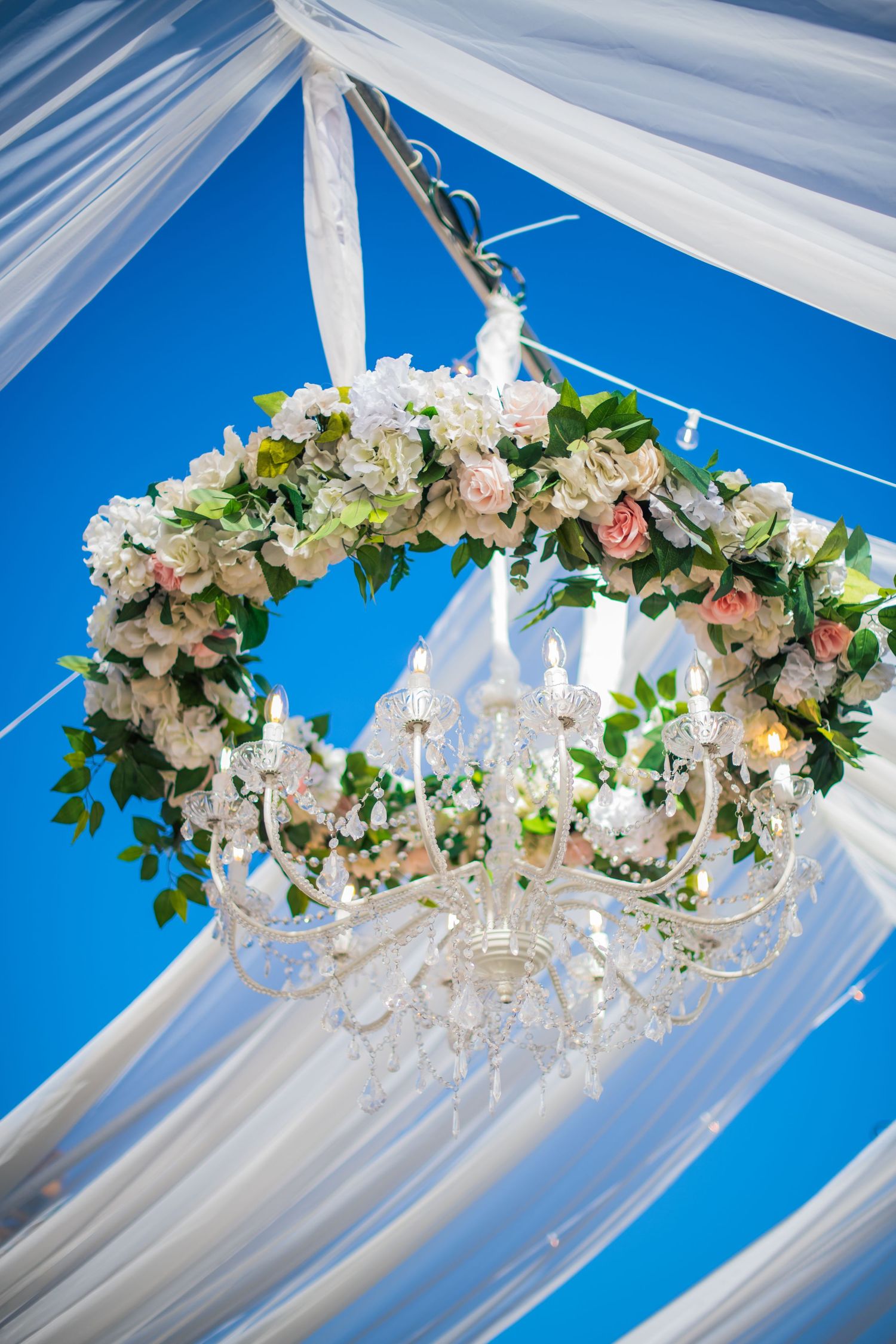 Elegant floral chandelier with white roses and crystals hangs from draped wedding tent ceiling.