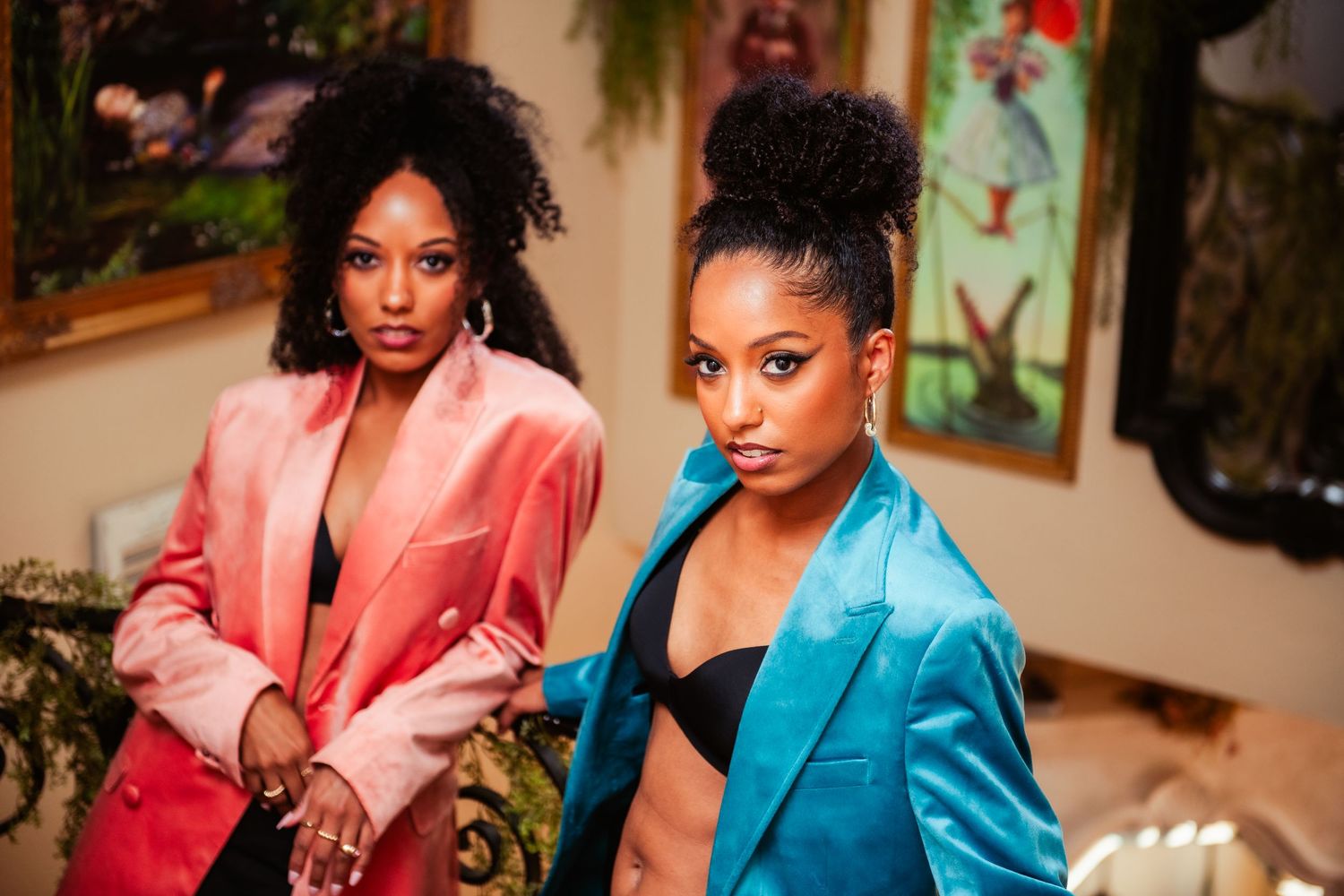 Two women pose in vibrant satin blazers, one in coral pink and one in turquoise blue, against an interior backdrop.