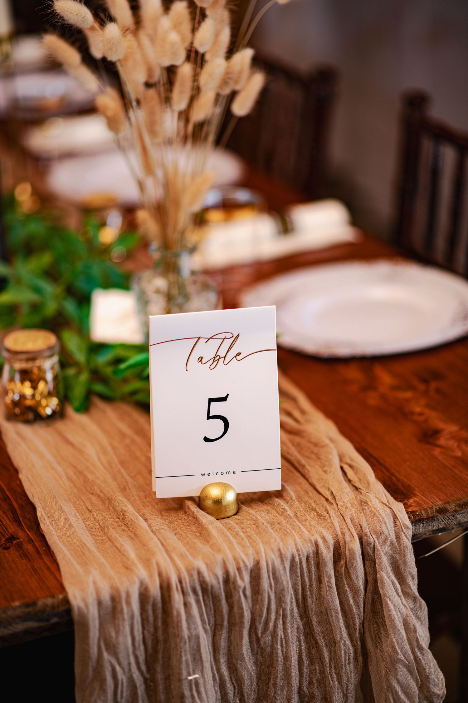Table number cards and place settings on rustic wooden tables with gold accents and greenery for an elegant wedding reception.