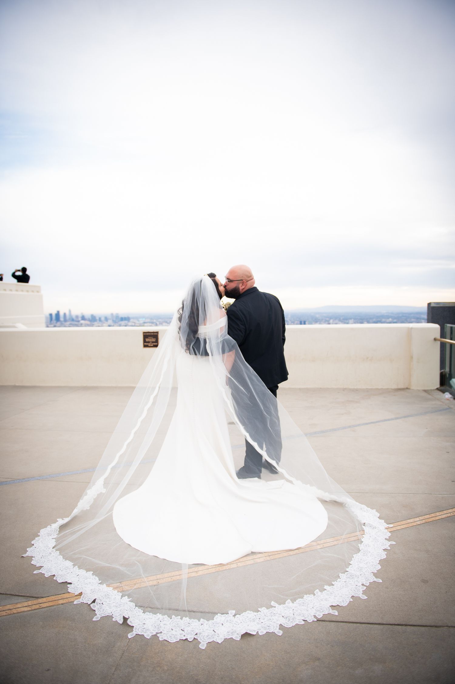 A wedding couple stands on a rooftop terrace with a dramatic cathedral-length lace veil flowing in the breeze.