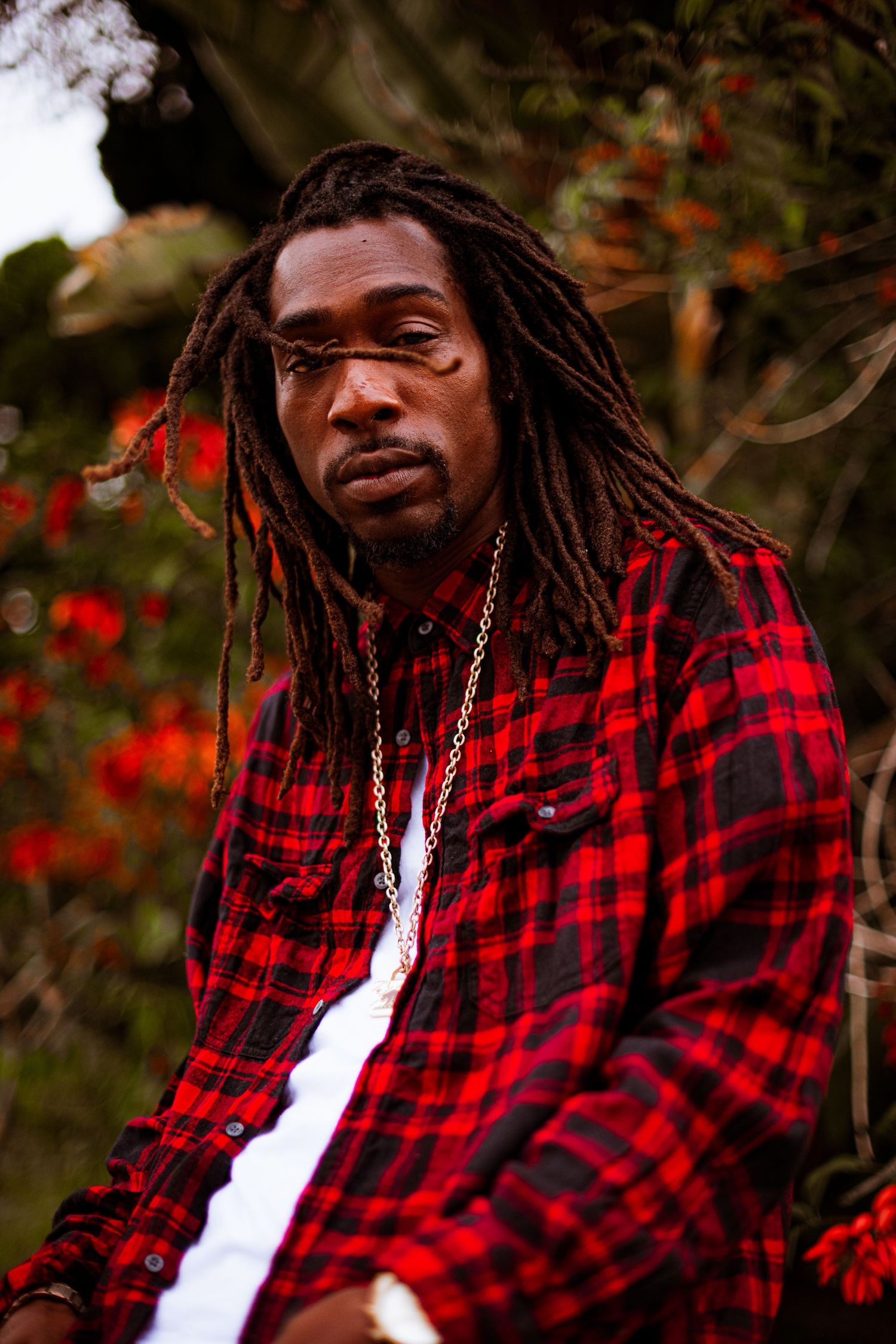 Person in red and black plaid flannel shirt with dreadlocks poses against autumn foliage background.