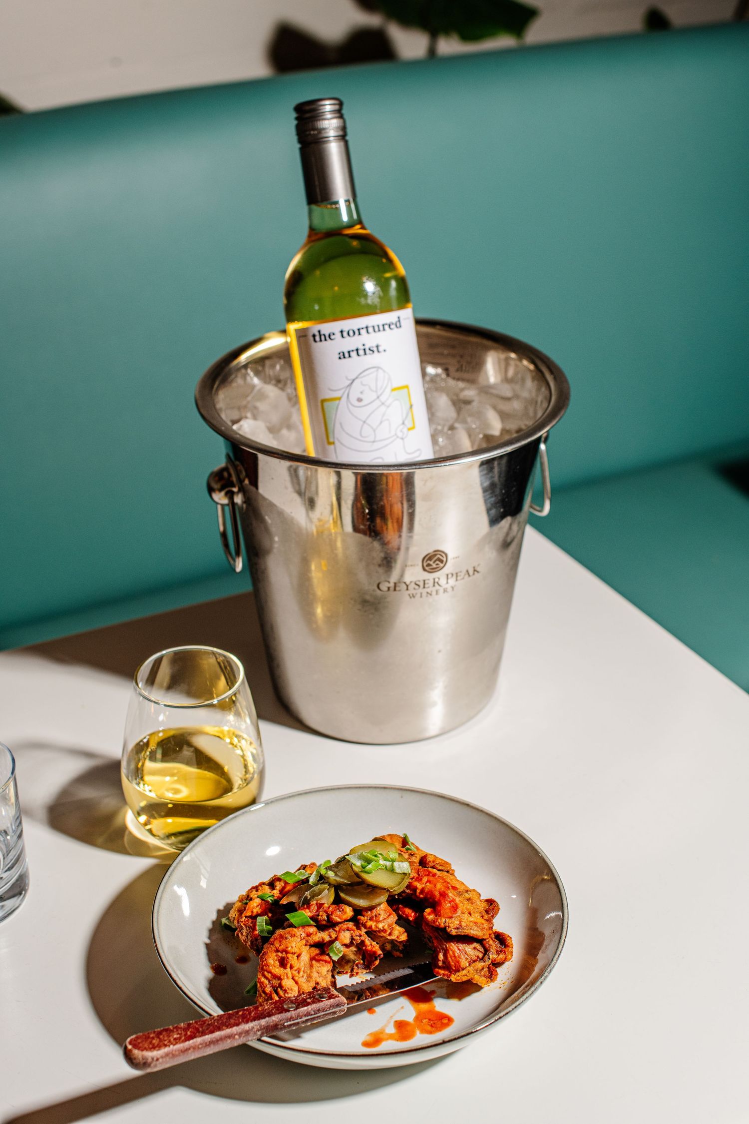 White wine chilling in silver ice bucket alongside spicy chicken dish on restaurant table.