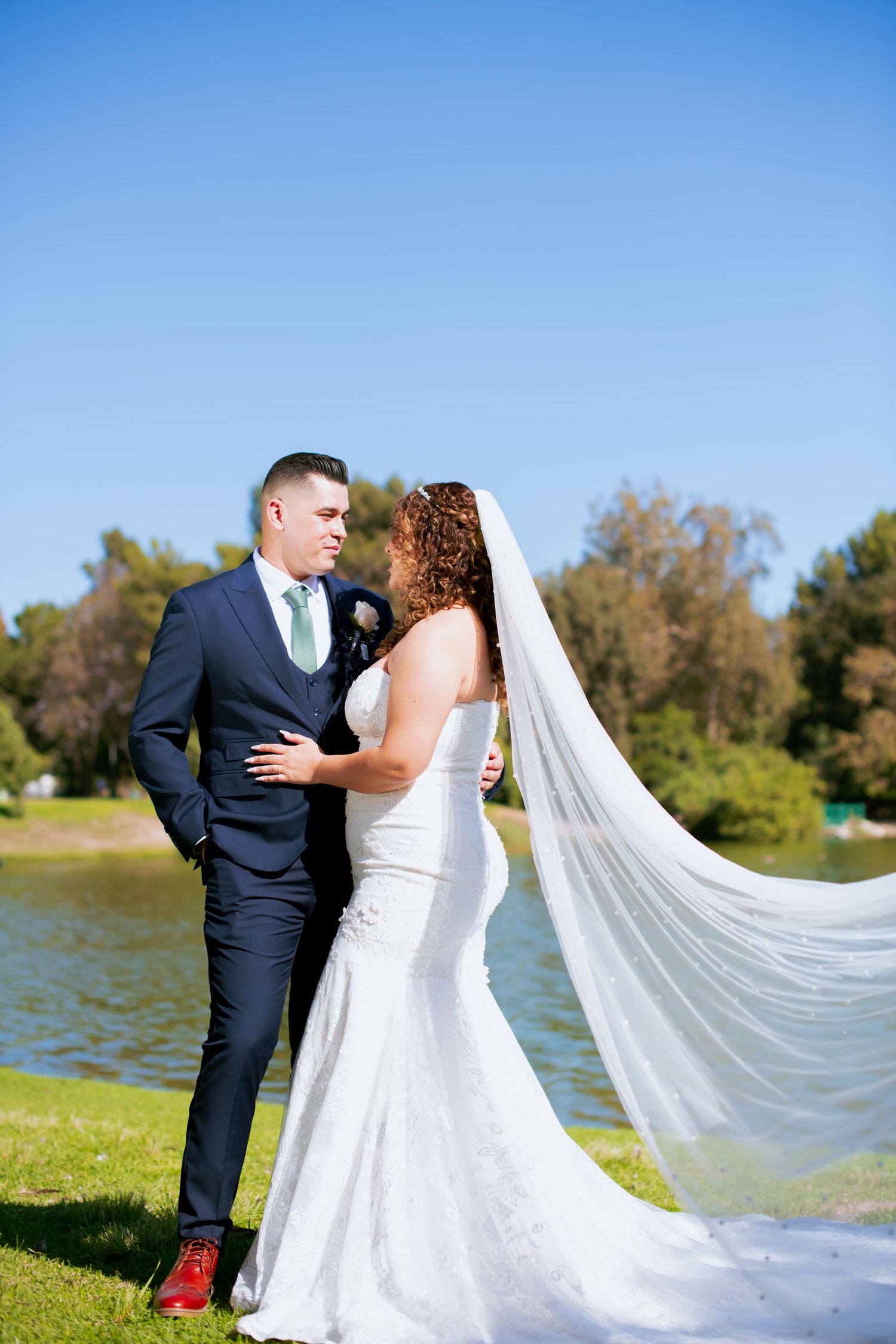 Wedding couple embracing near lake with dramatic flowing veil and clear blue sky.
