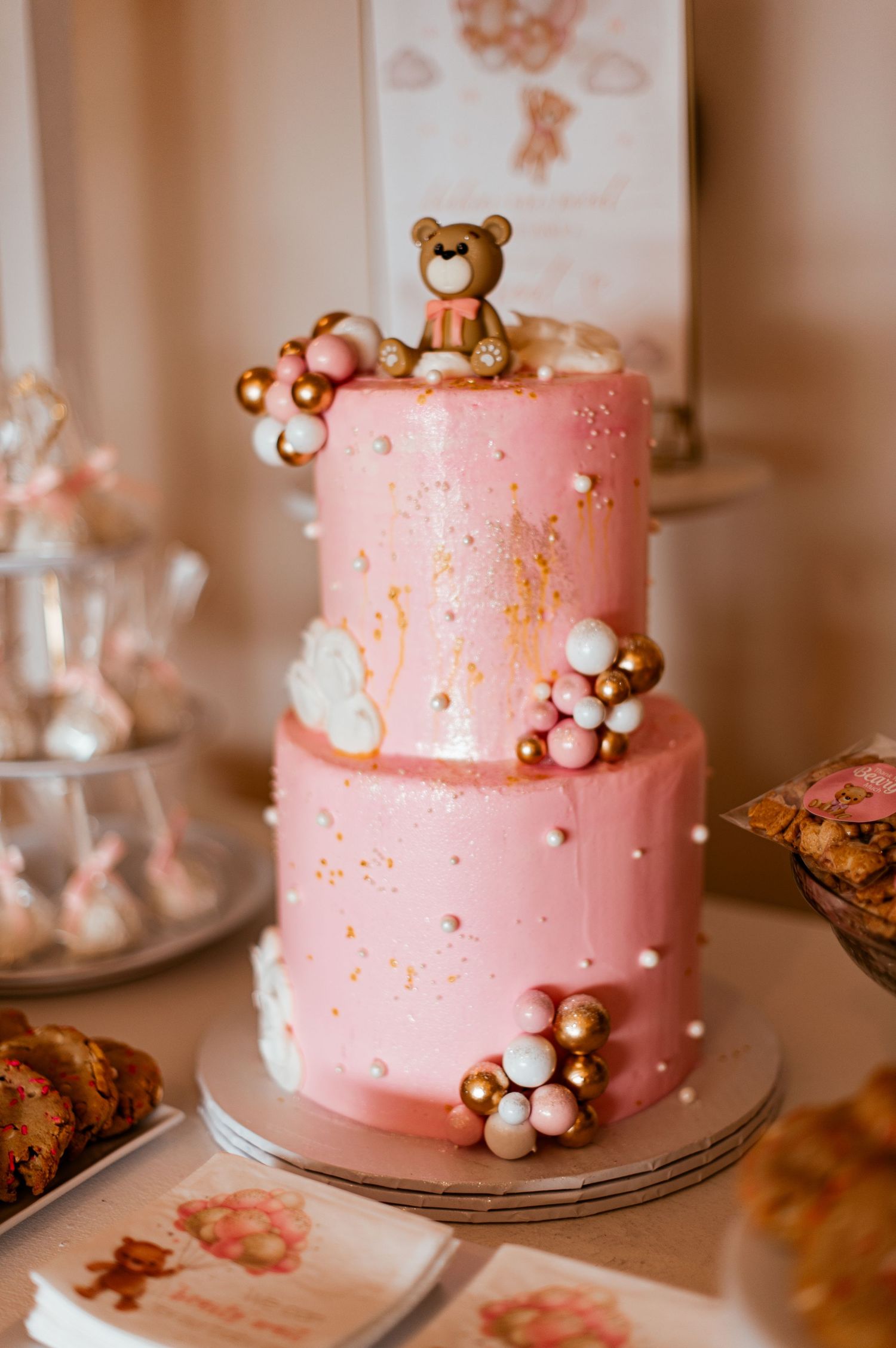 Two-tiered pink baby shower cake decorated with gold accents and teddy bear topper.