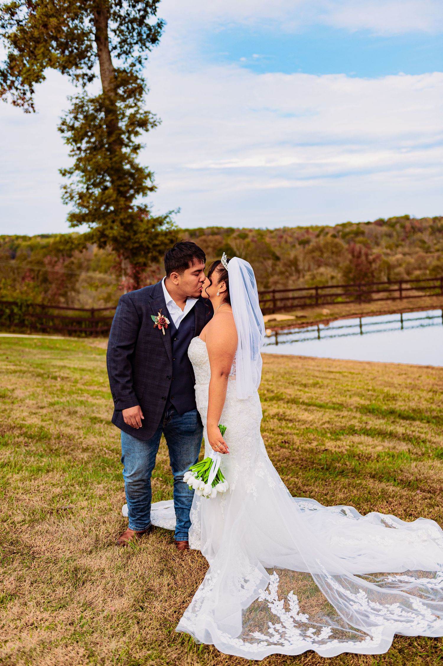 Bride in flowing white lace dress poses with groom in scenic countryside setting with autumn trees.