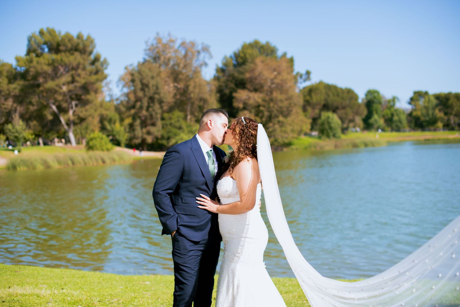 Romantic lakeside wedding photo with a flowing veil against blue water and green trees.
