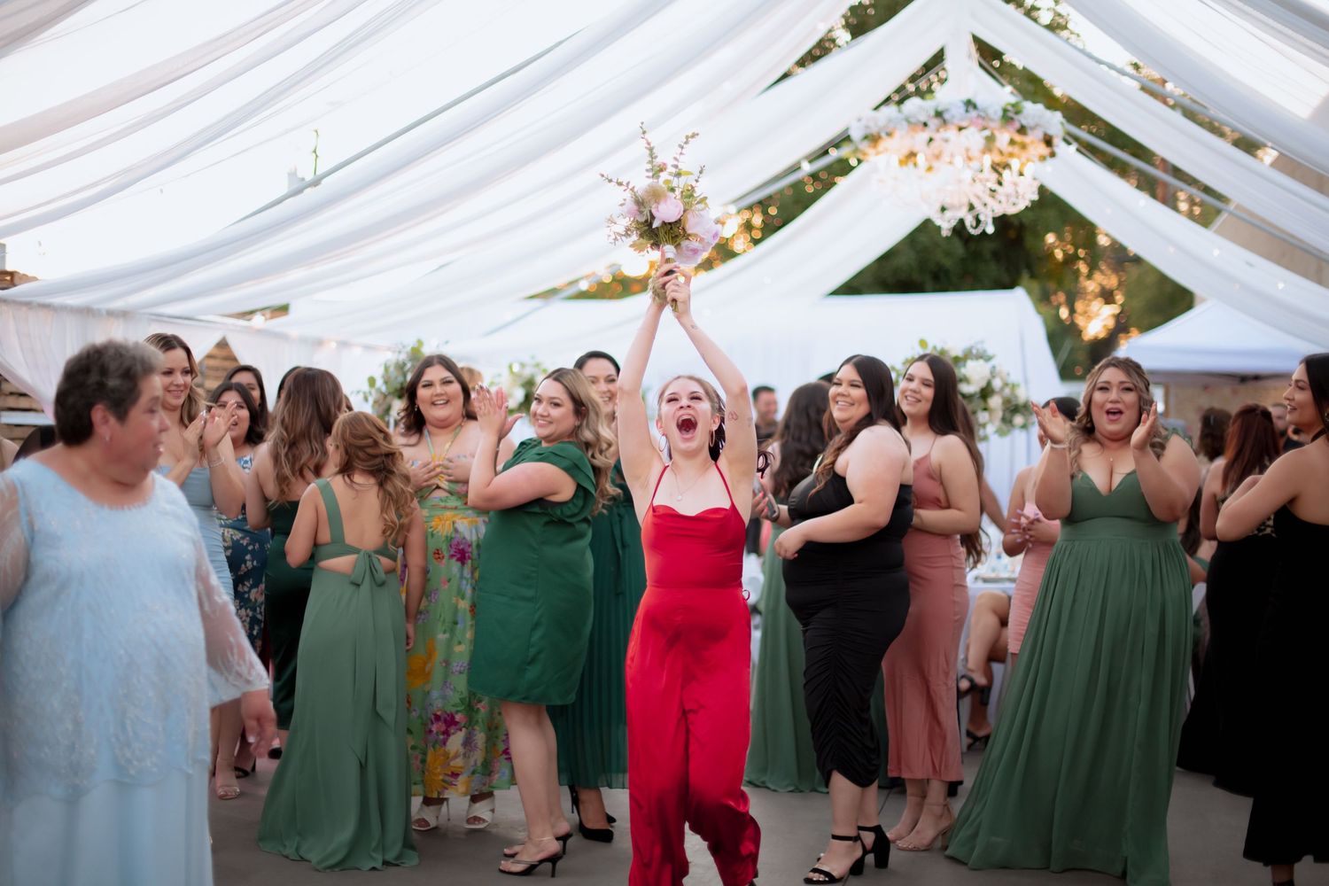 Wedding party members in green and red dresses celebrating on the dance floor under white tent.