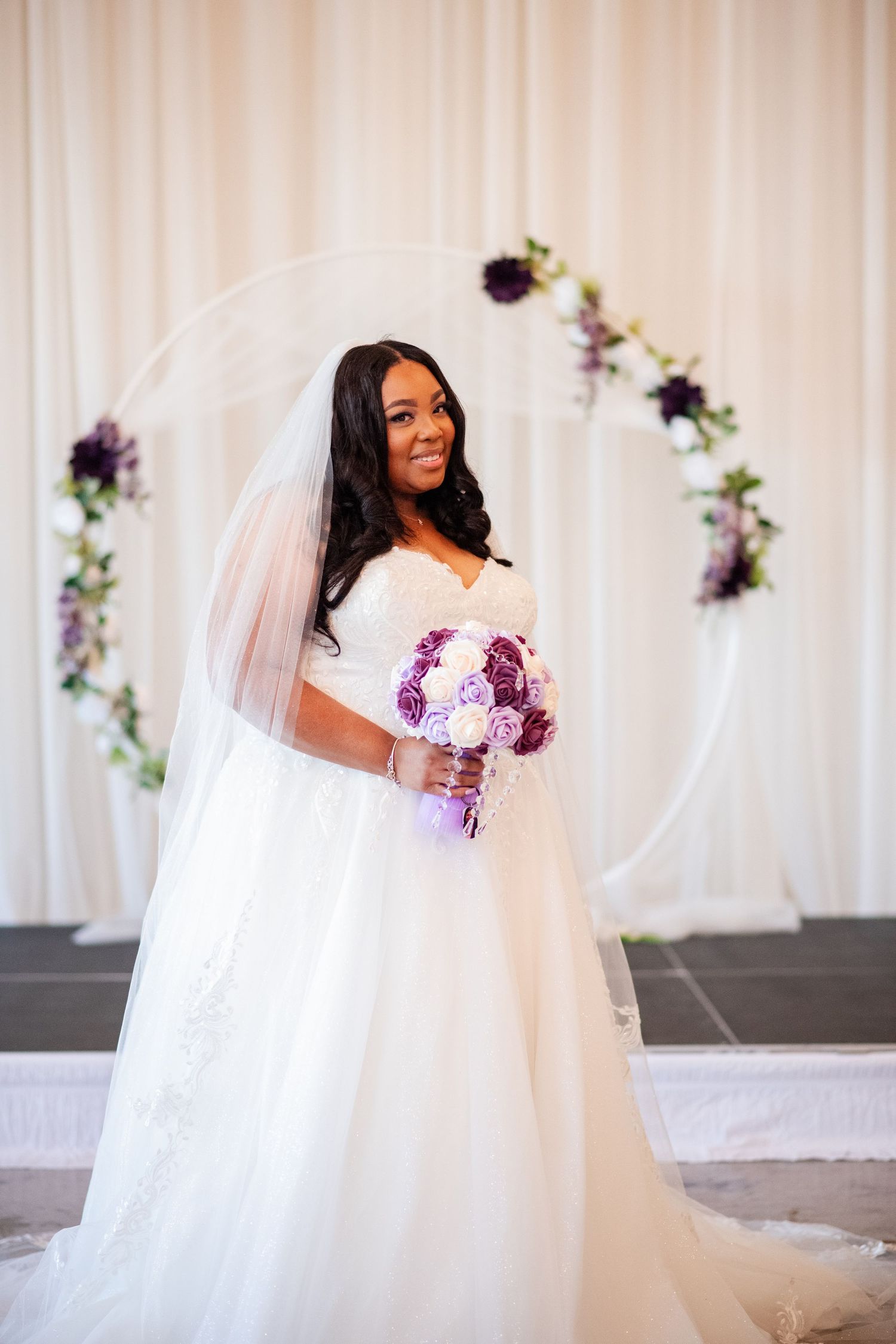 Bride in white wedding gown holding purple and white rose bouquet against light backdrop.