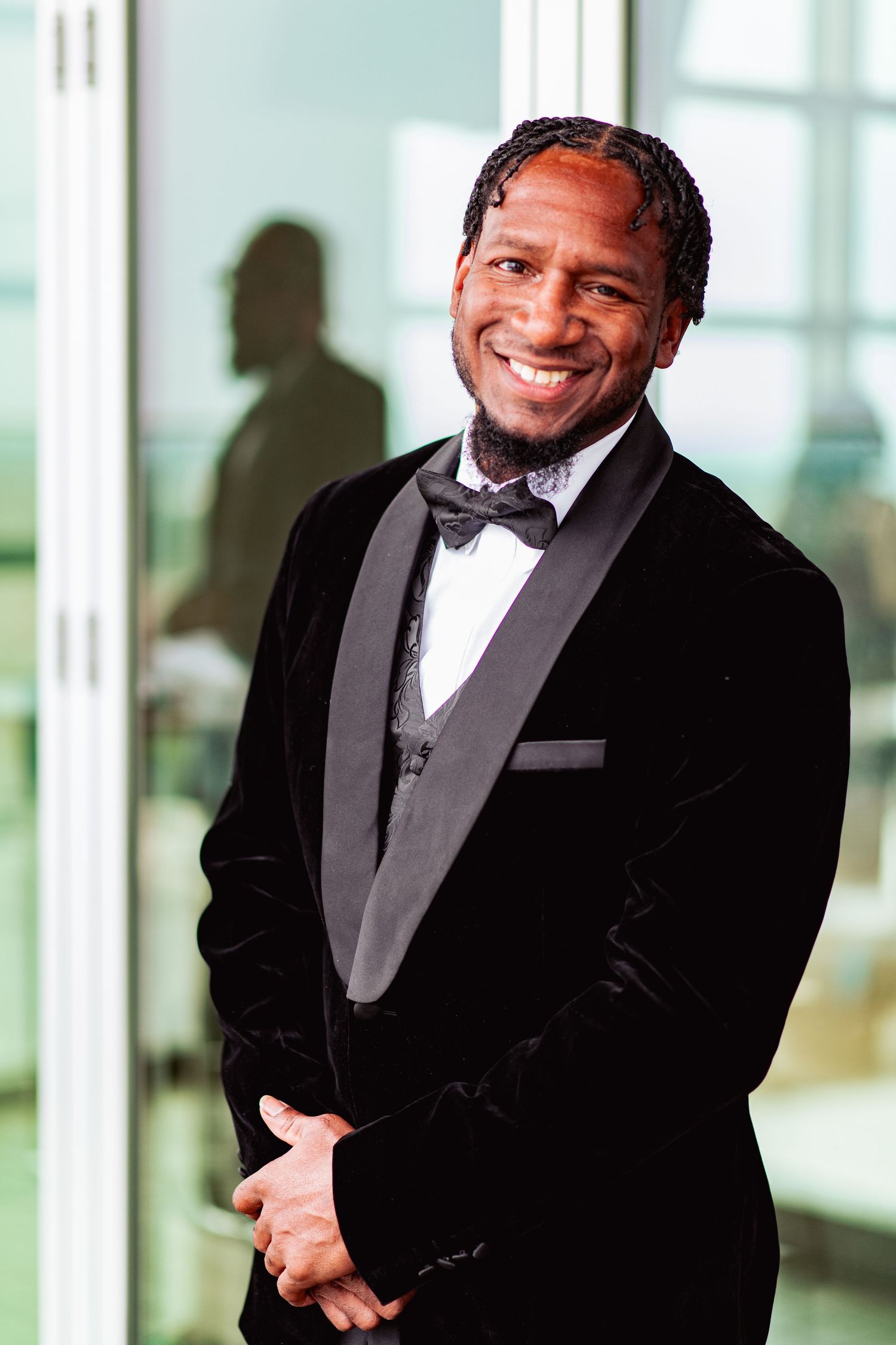 A person in a black tuxedo and bowtie smiles warmly in a bright indoor setting.