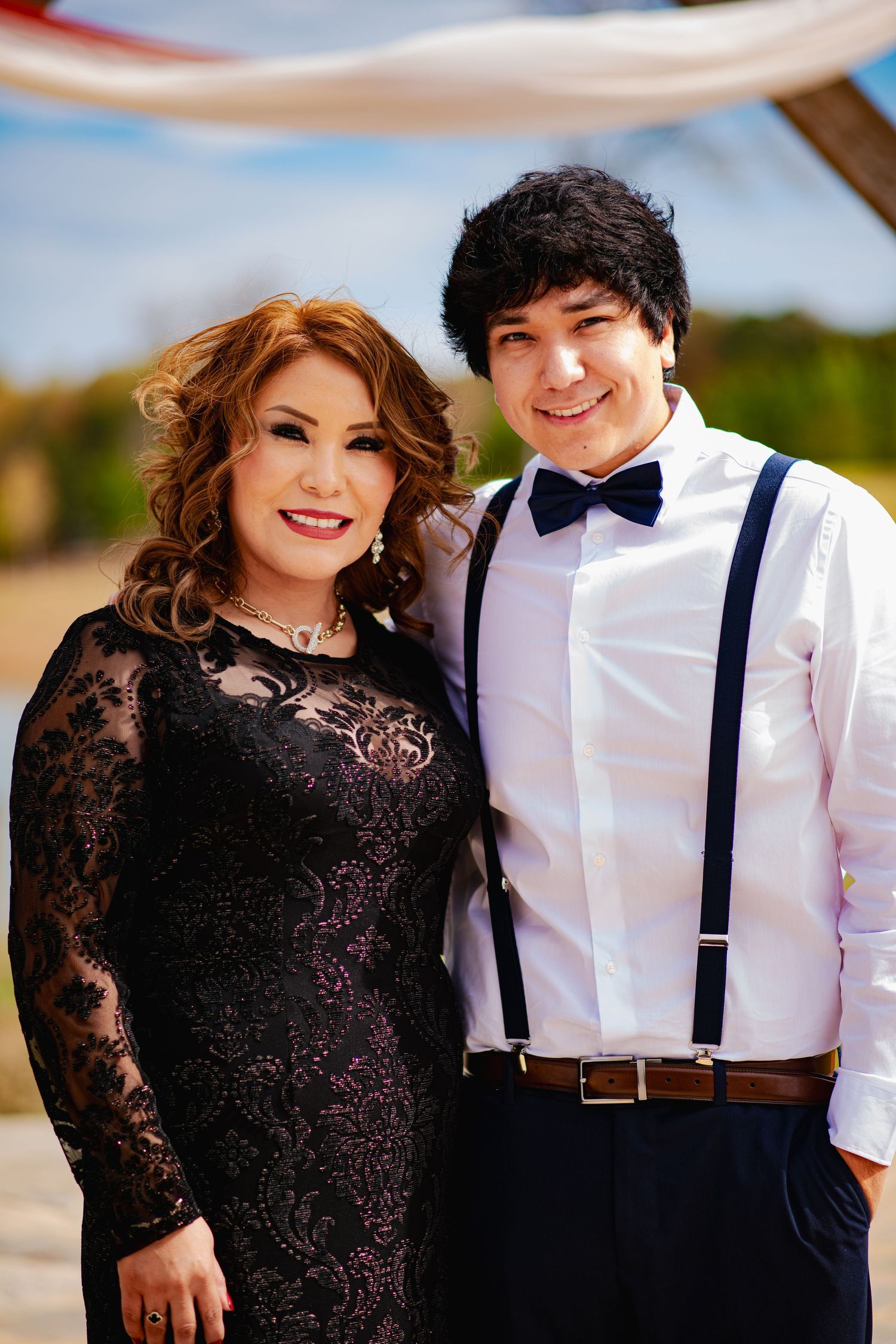 A couple poses together at a formal event, with the woman in a black lace dress and the man in suspenders and bow tie.