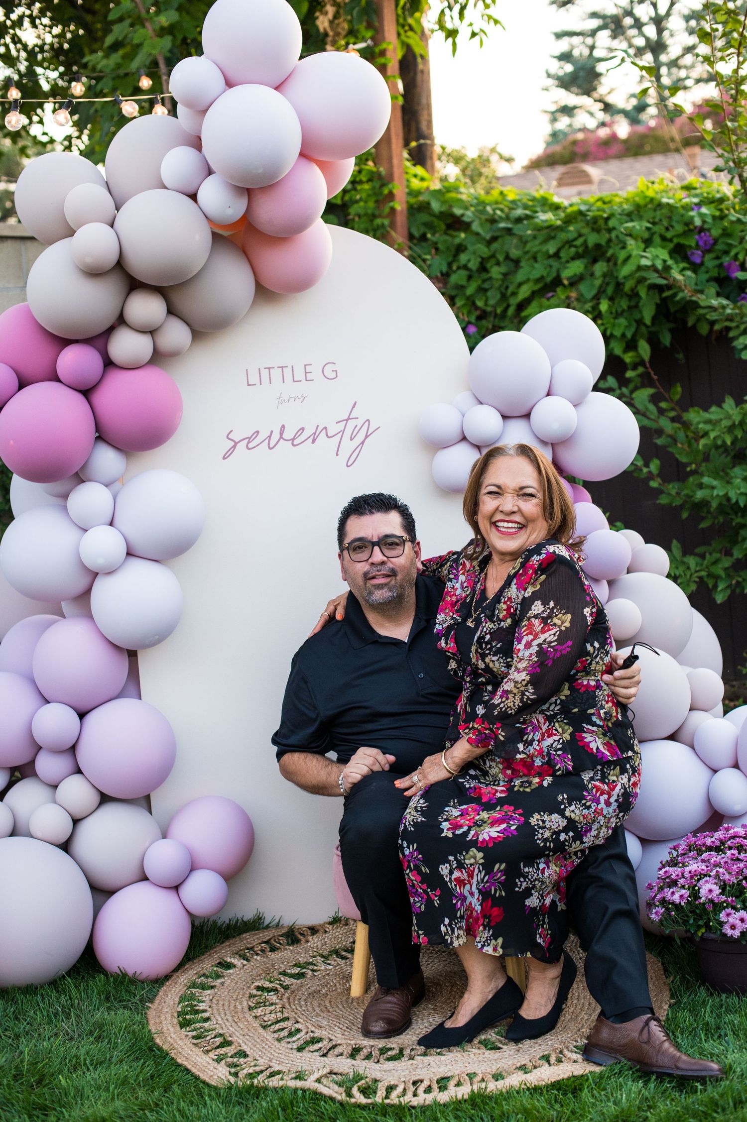 A celebratory setup with pink and white balloon arch decorations and a white circular backdrop in an outdoor garden setting.