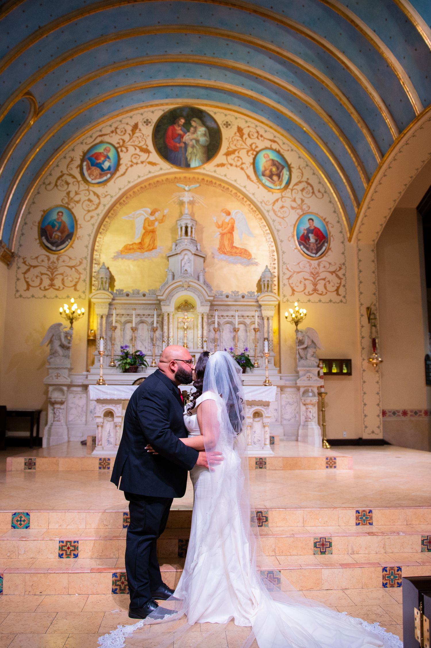 A couple shares an intimate moment at the altar of an ornate church with blue and gold painted ceiling murals.