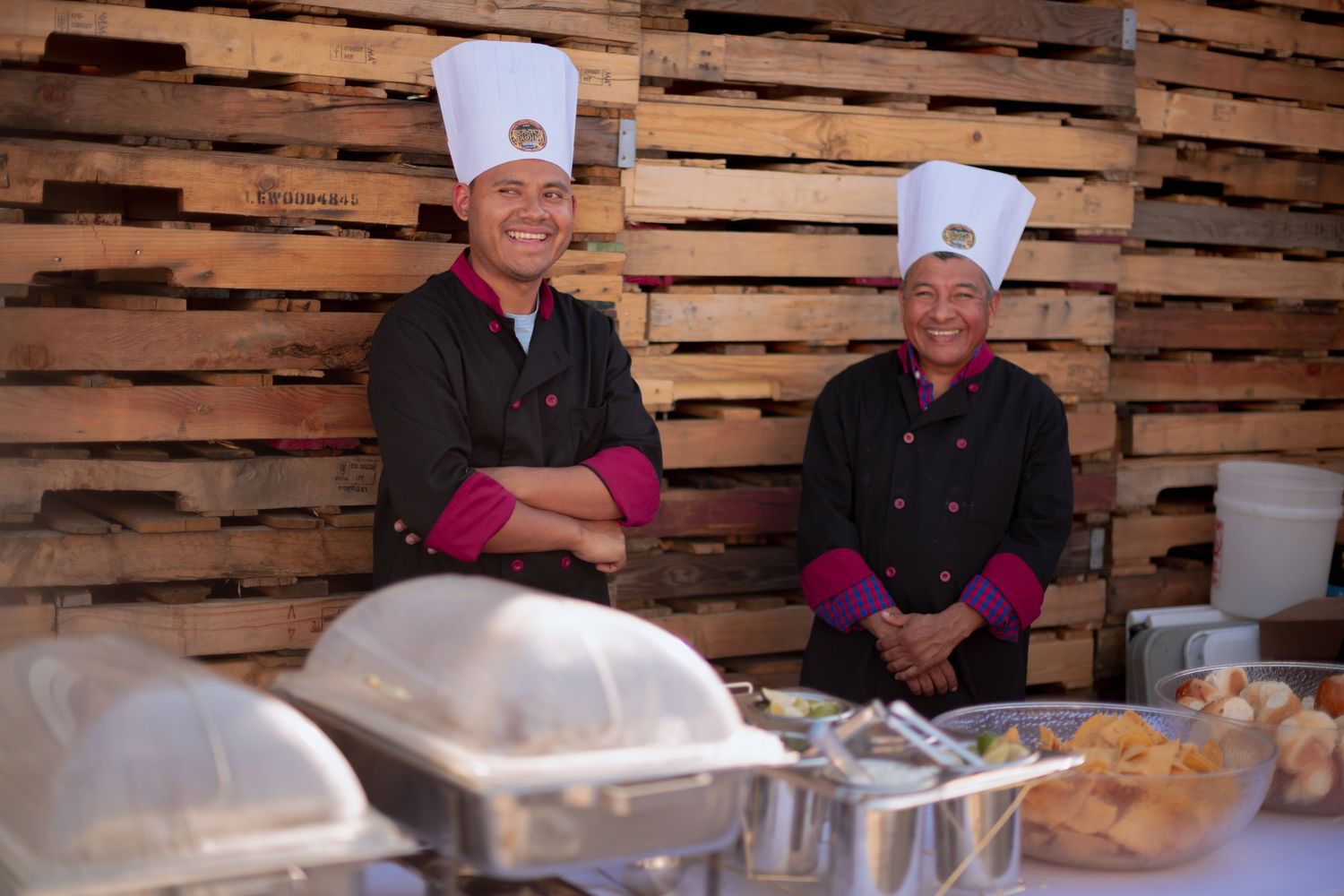 Chefs in white uniforms prepare food at a traditional buffet line against rustic wooden wall decor.