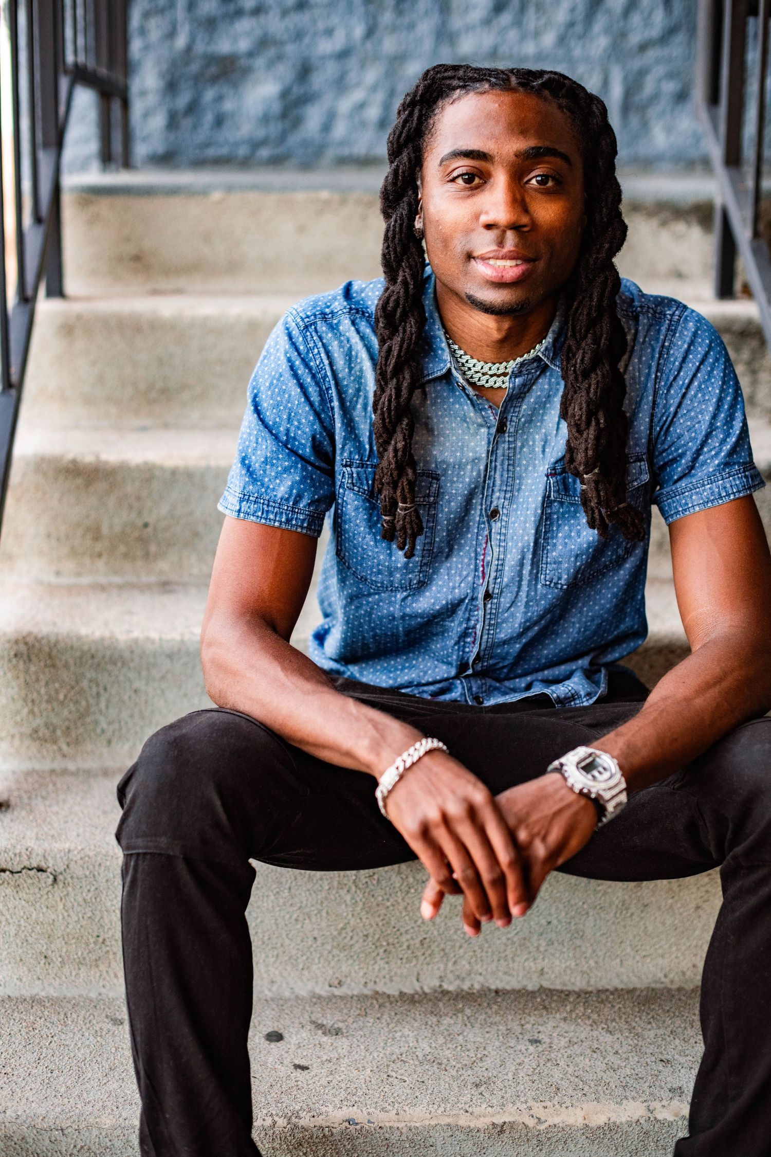 Person sitting on concrete steps wearing denim shirt and dark pants in casual outdoor photoshoot.