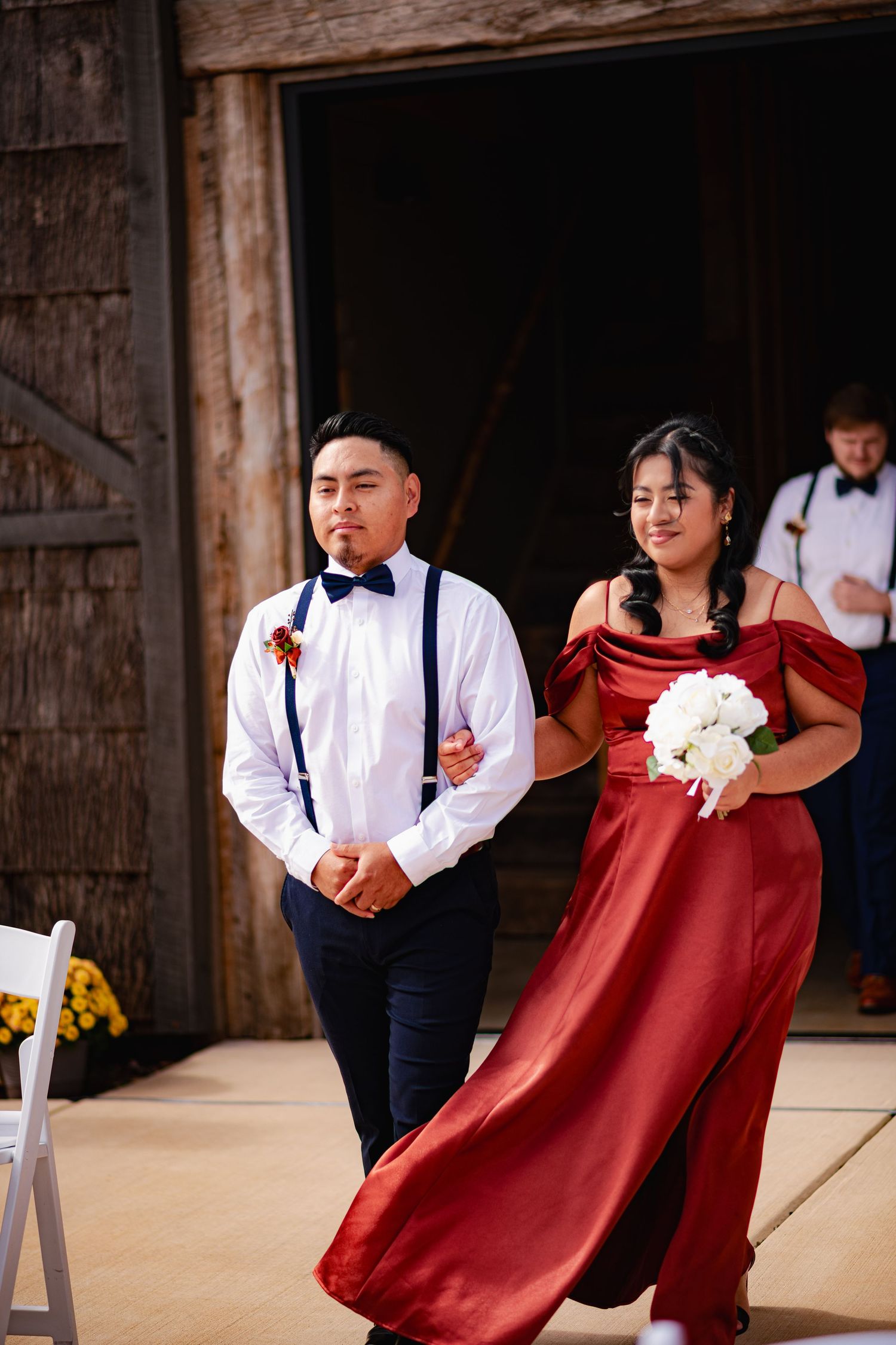 Wedding party members in burgundy dresses and navy suits dance playfully in a rustic barn setting.