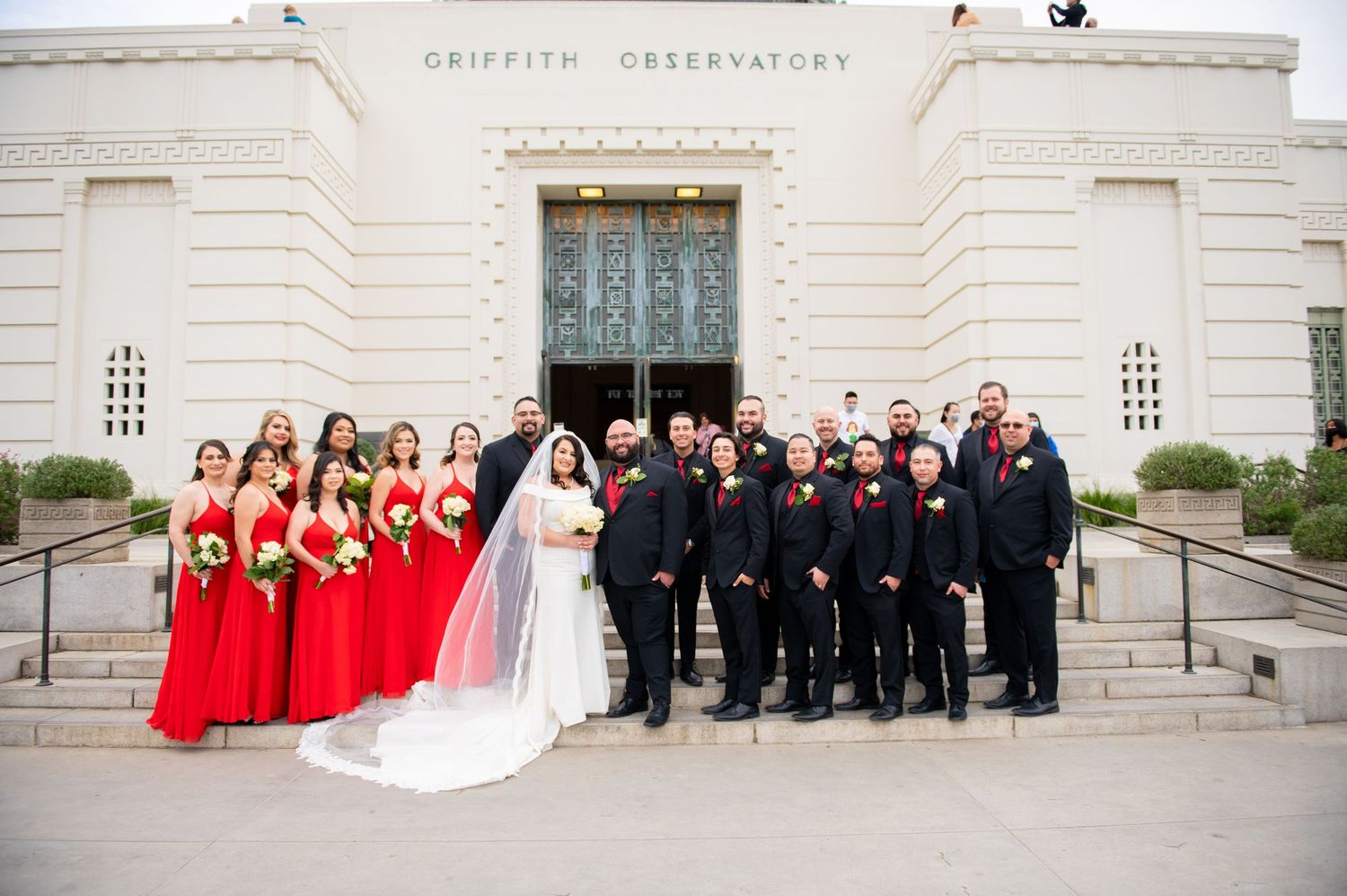 Wedding party members pose on grand stone steps of Griffith Observatory in formal attire.