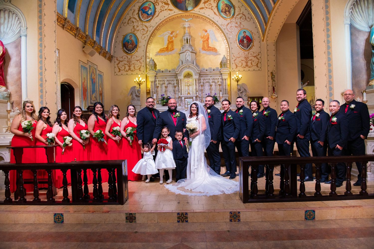 Wedding party in formal red and black attire pose in ornate church sanctuary with elegant altar and religious artwork.