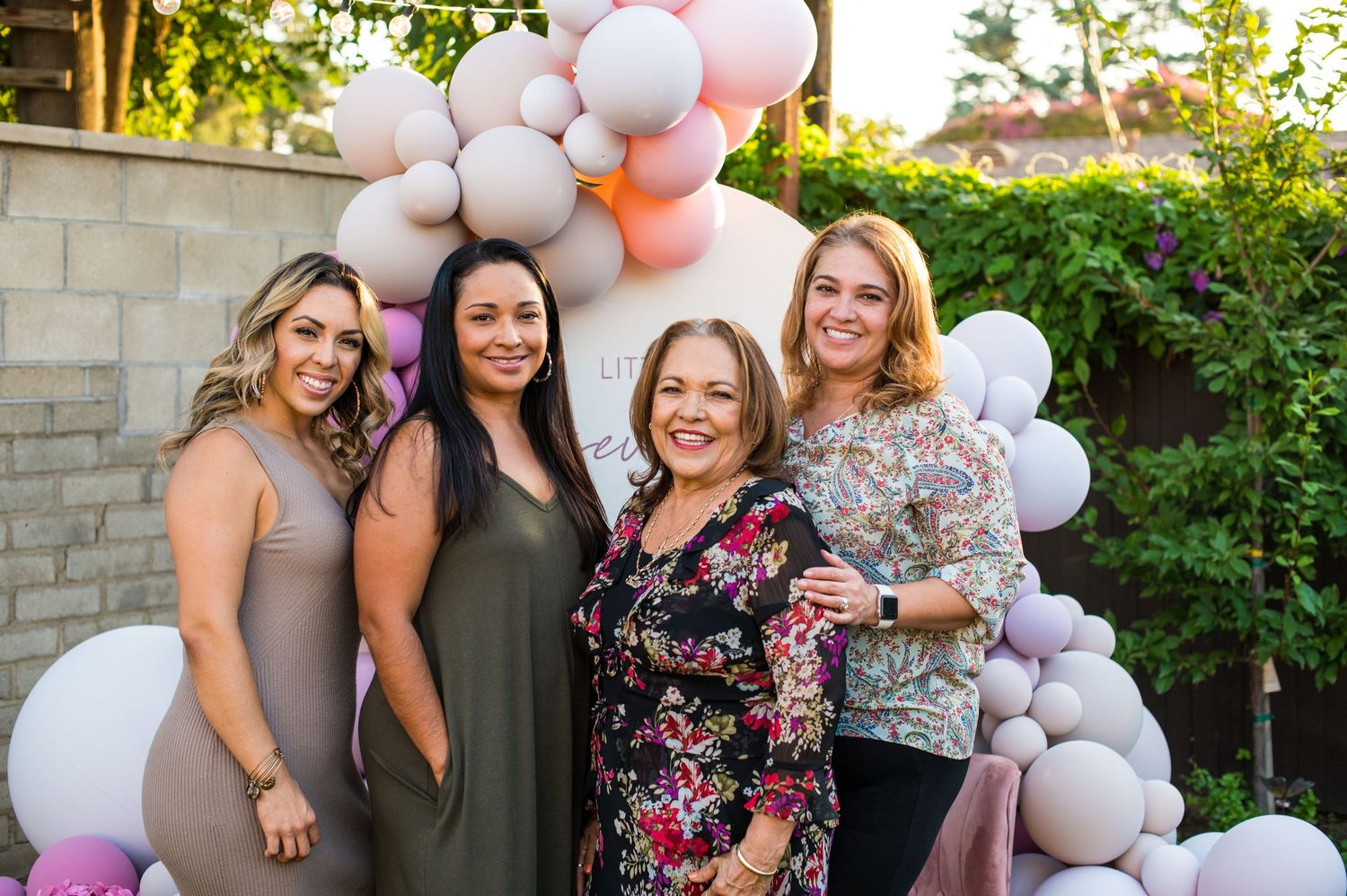 A group poses together at an outdoor celebration decorated with pink and white balloon garlands and greenery.