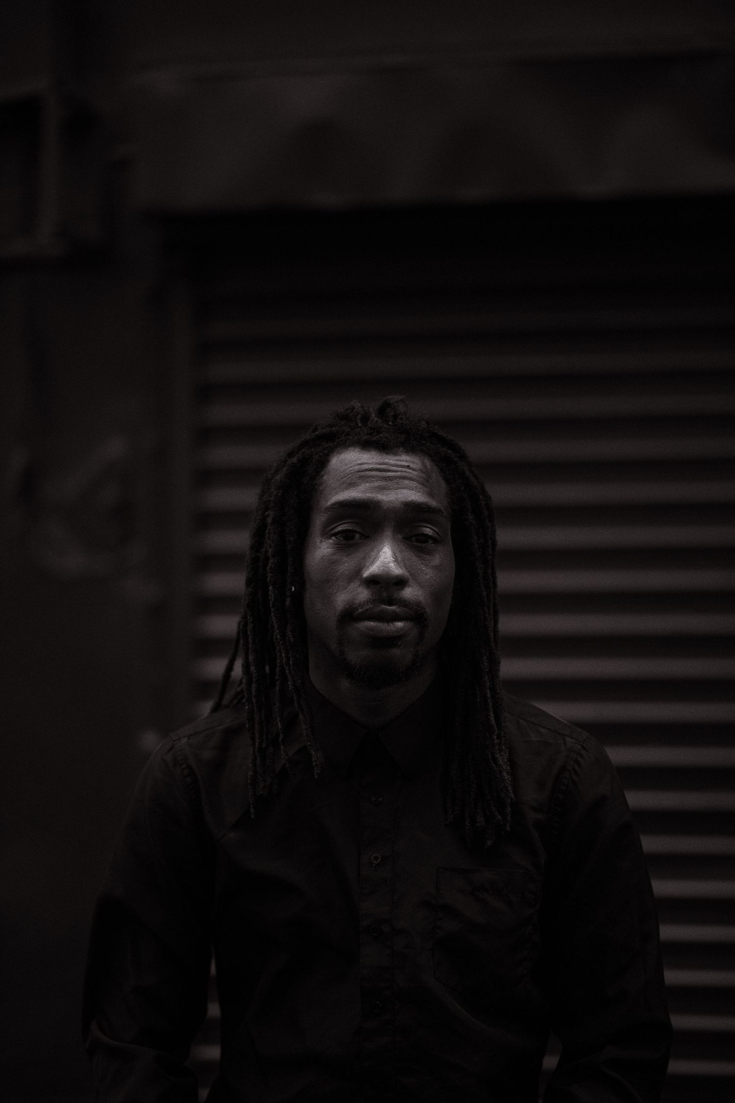 Moody black and white portrait of person with dreadlocks wearing dark clothing against industrial background.