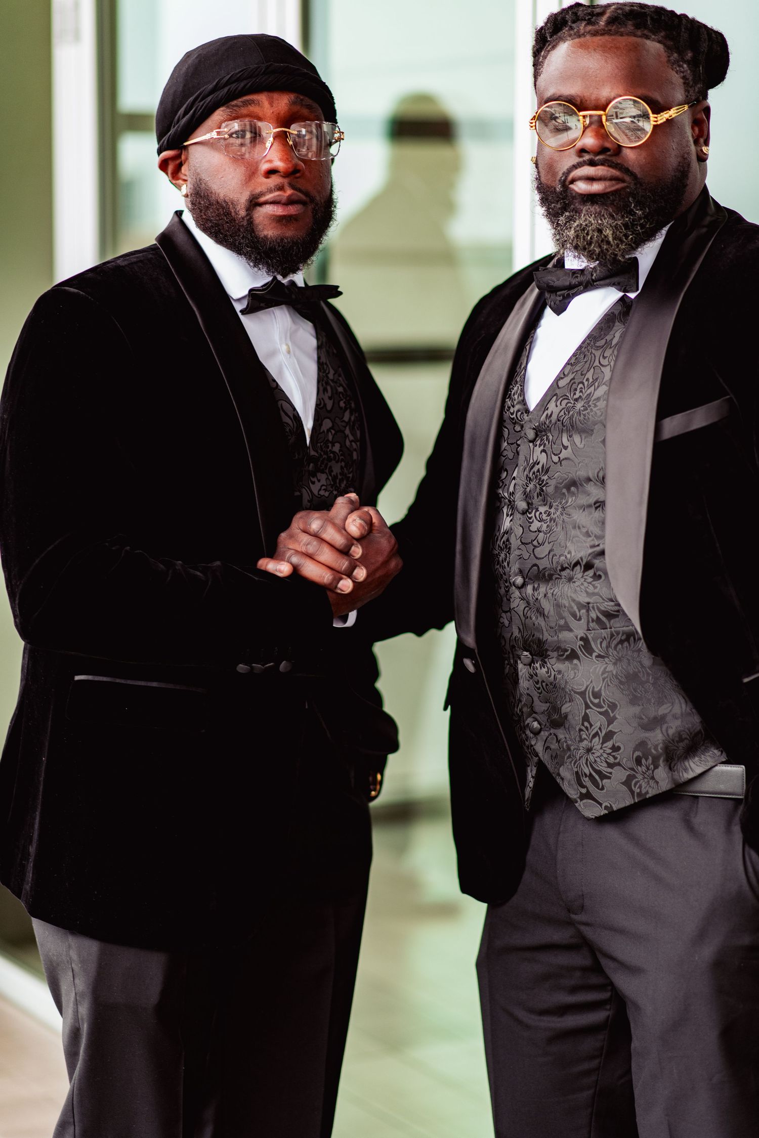 Two men in formal black suits and vests pose together in an elegant indoor setting.
