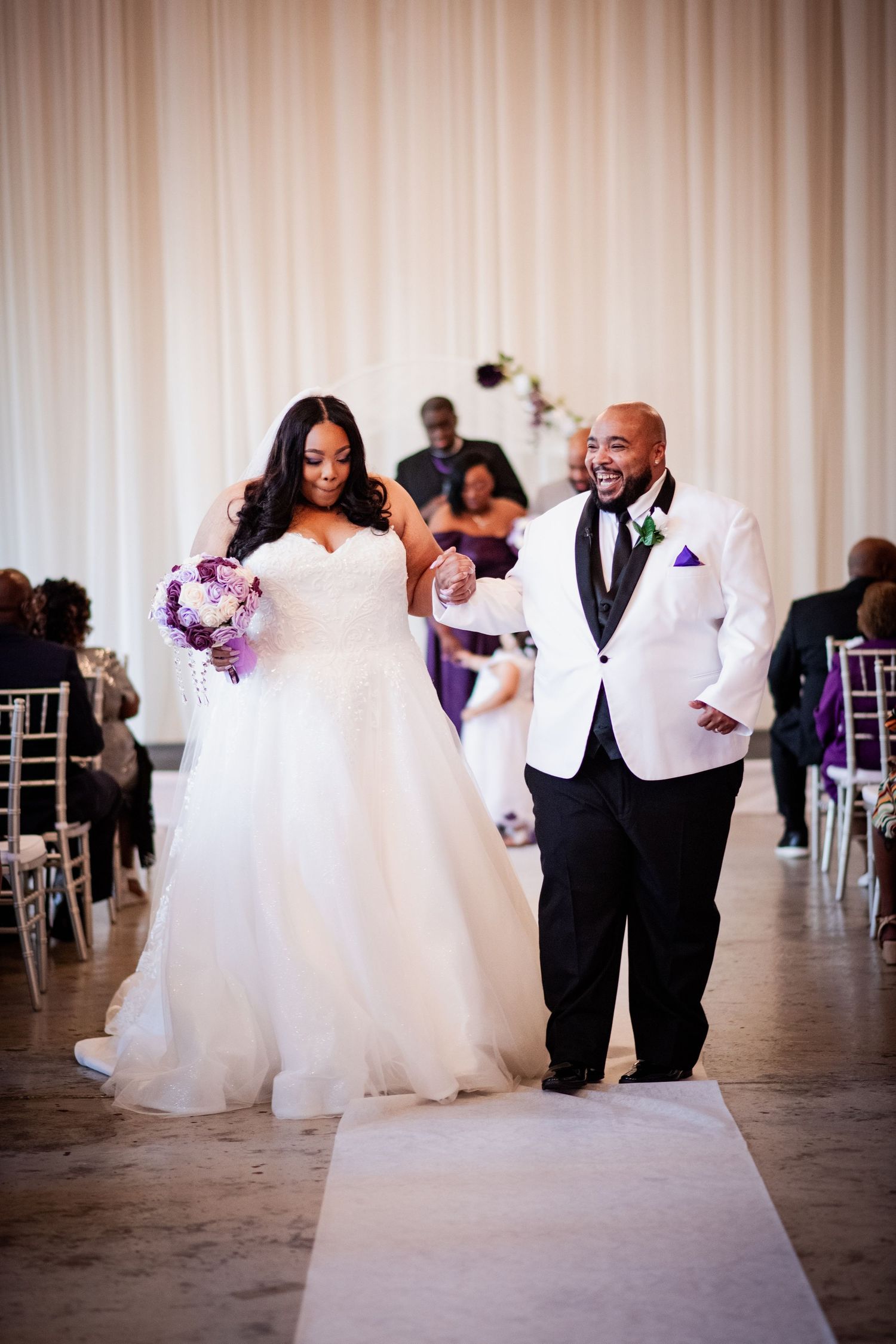 Wedding party members in white and purple attire dancing down an aisle.