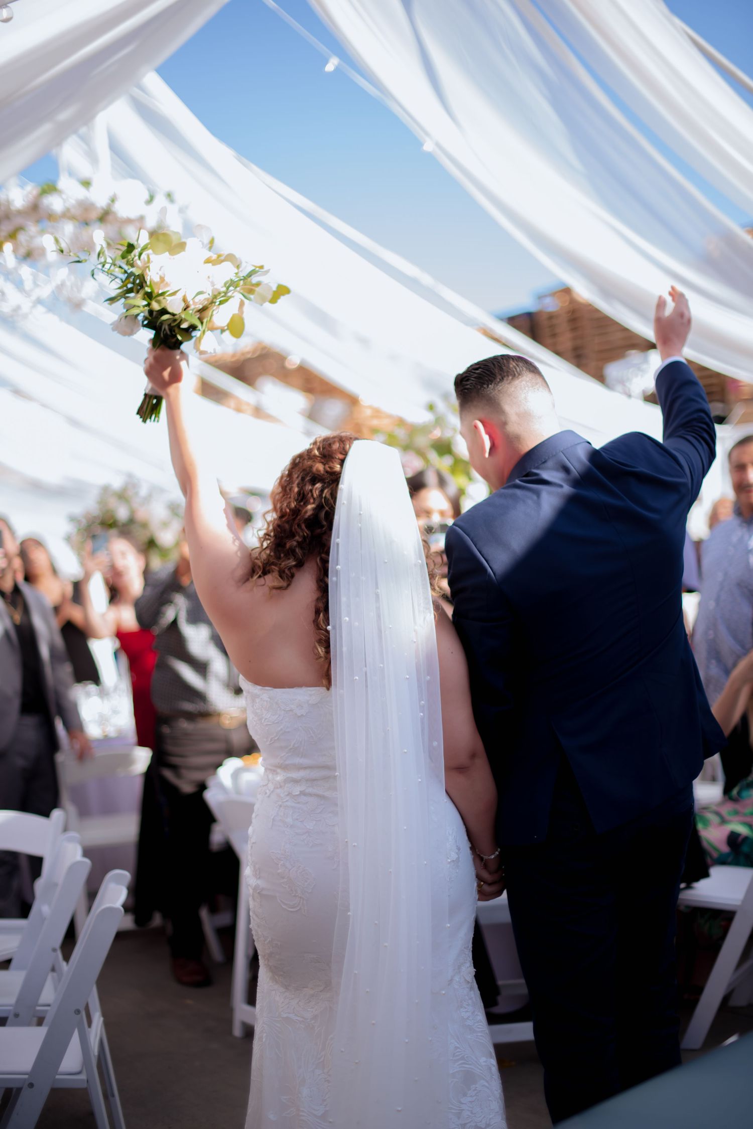 Wedding guests and newlyweds celebrating under white draped fabric at an outdoor reception.