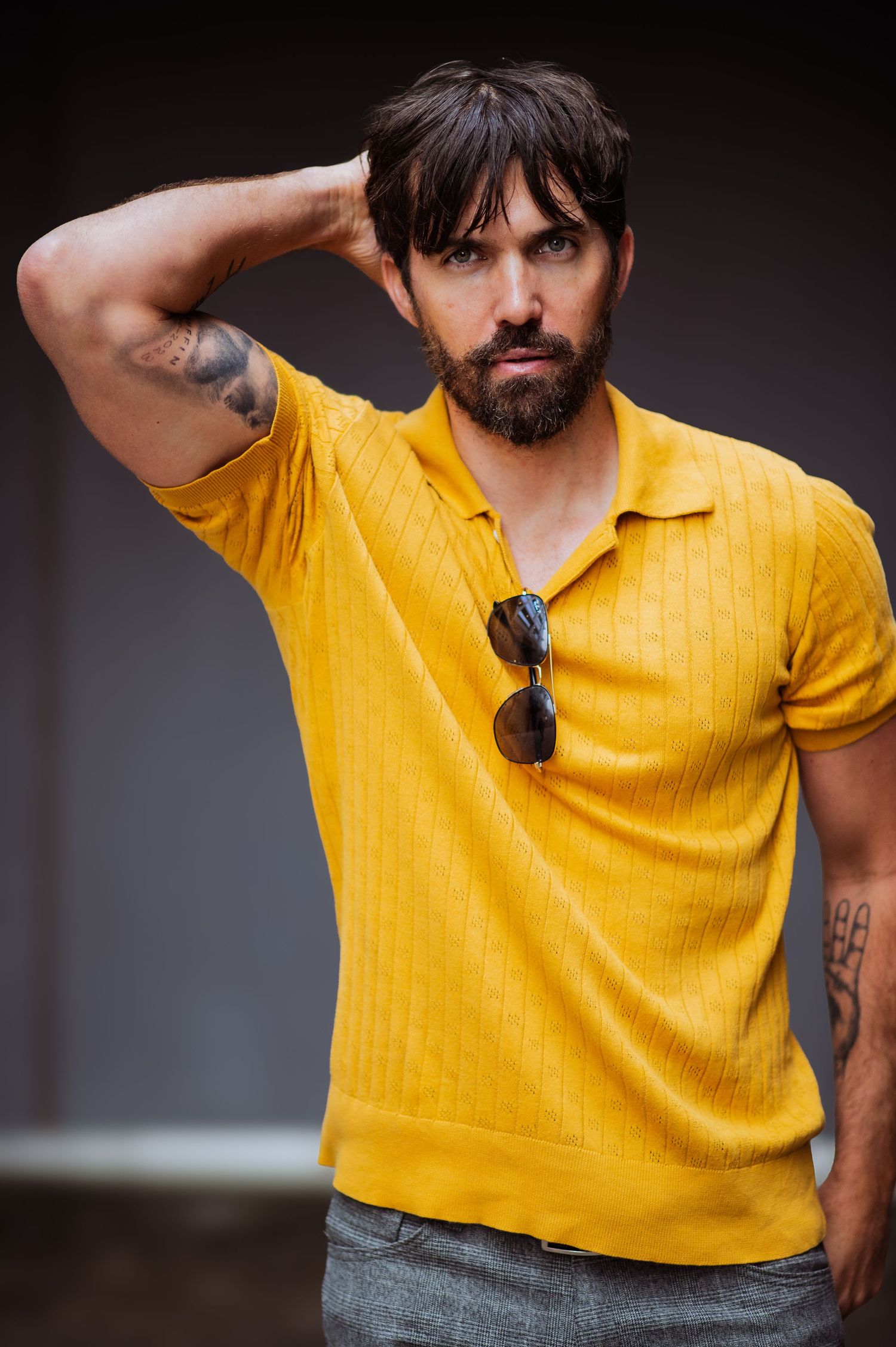 Athletic figure in yellow knit polo shirt with tattoos and sunglasses strikes casual pose.