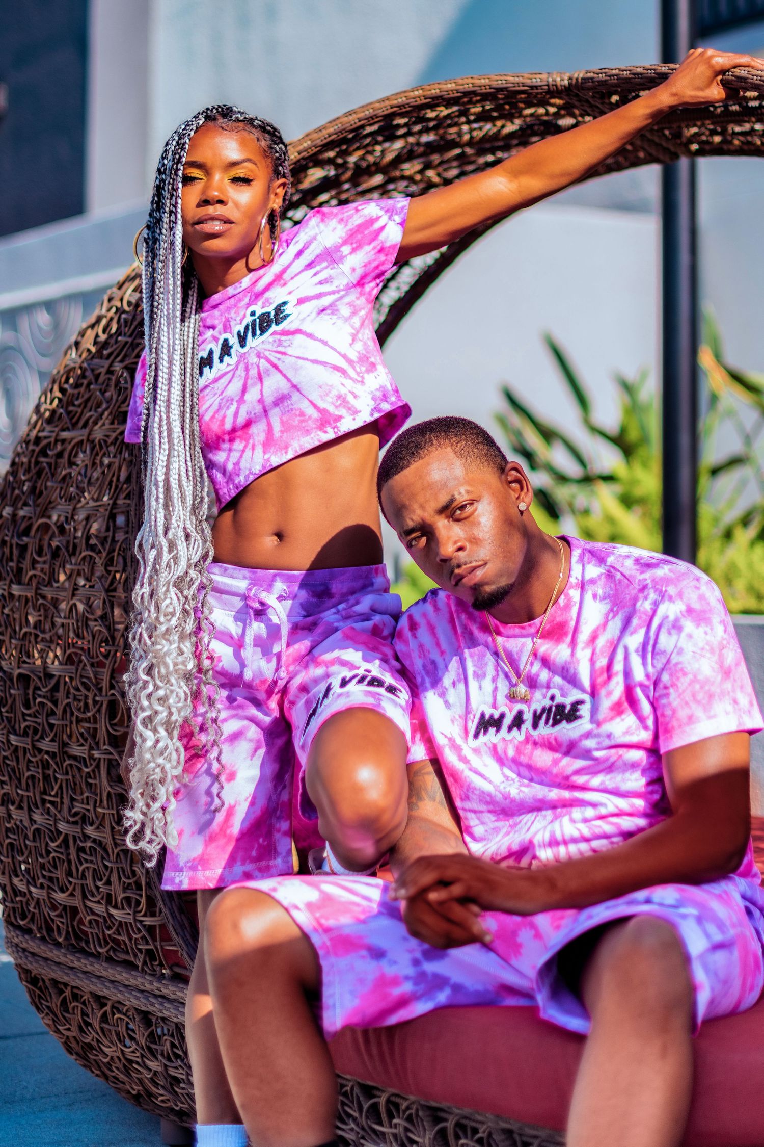 Models wearing pink tie-dye shirts pose on outdoor furniture during fashion photoshoot.