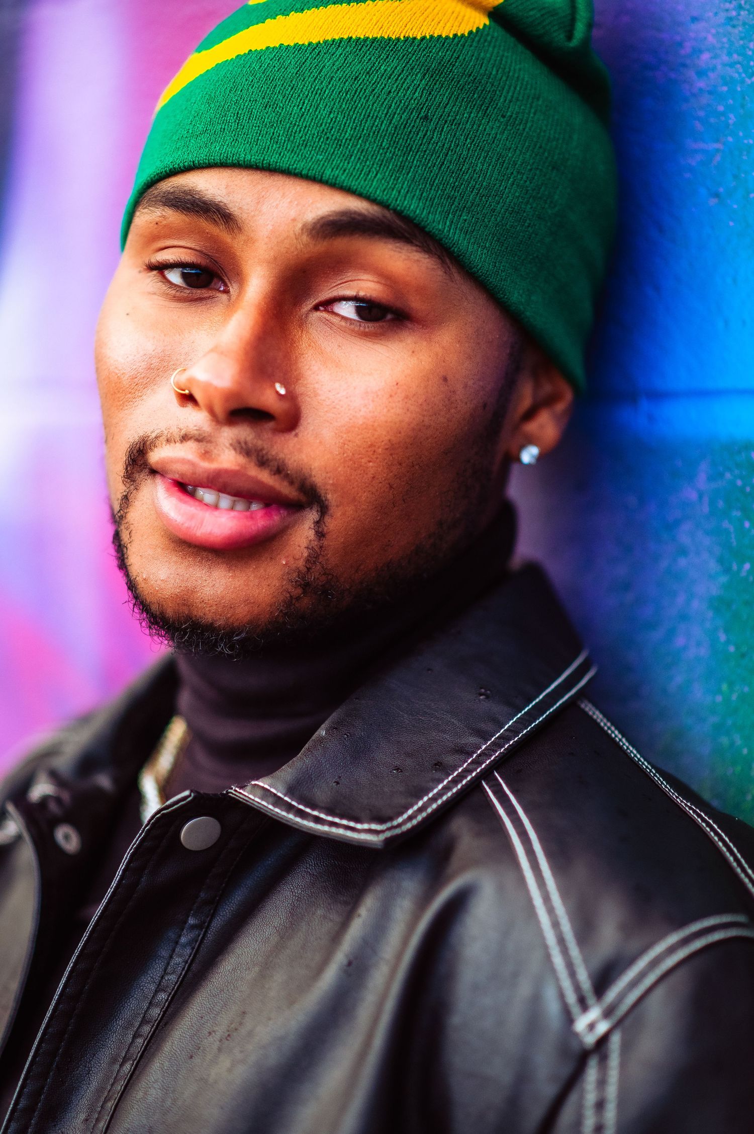 Person in green beanie and black jacket poses against colorful neon-lit background.