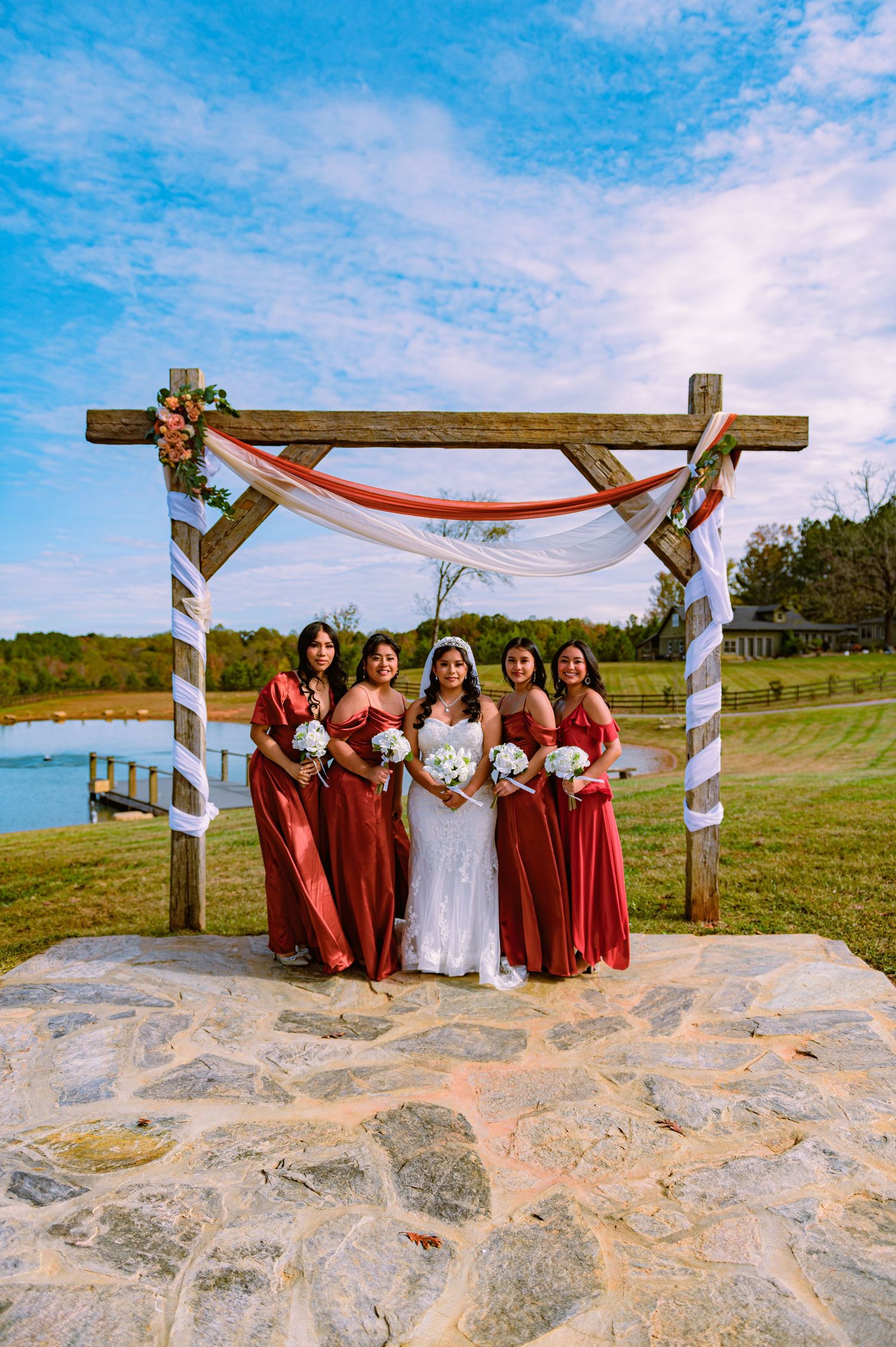 Wedding party in burgundy dresses poses under a wooden arch decorated with fabric on a stone platform by a lake.