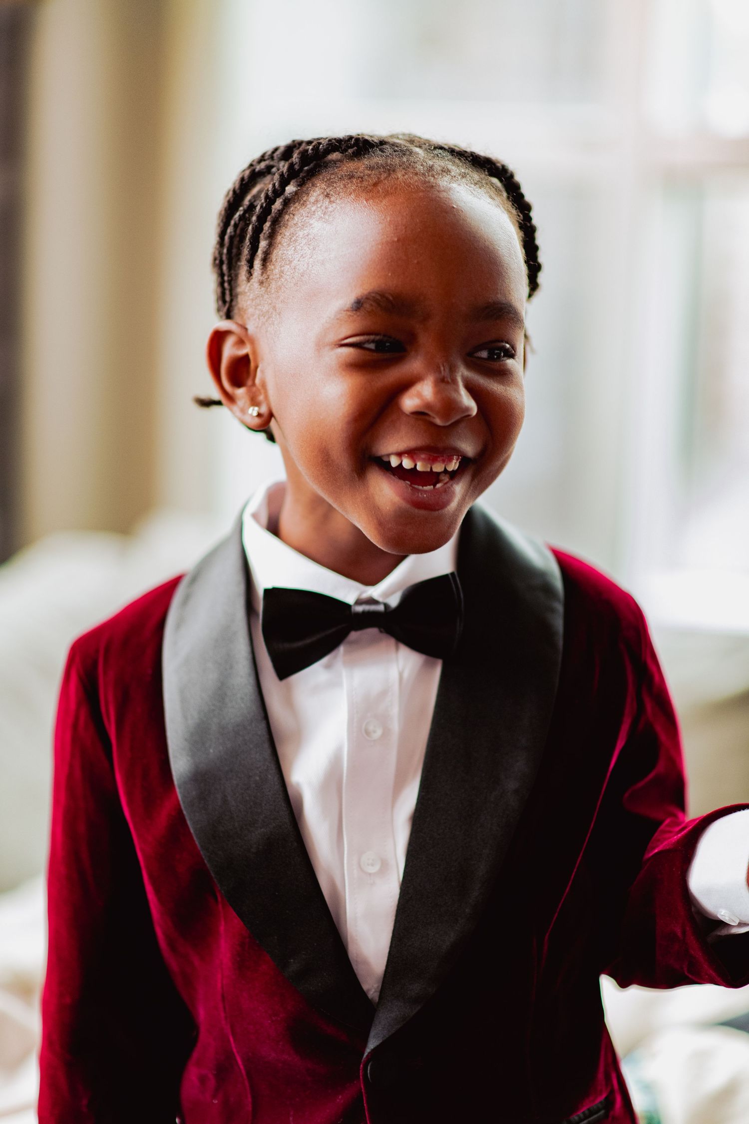 Young child wearing a red velvet jacket and black bow tie smiles brightly at the camera.