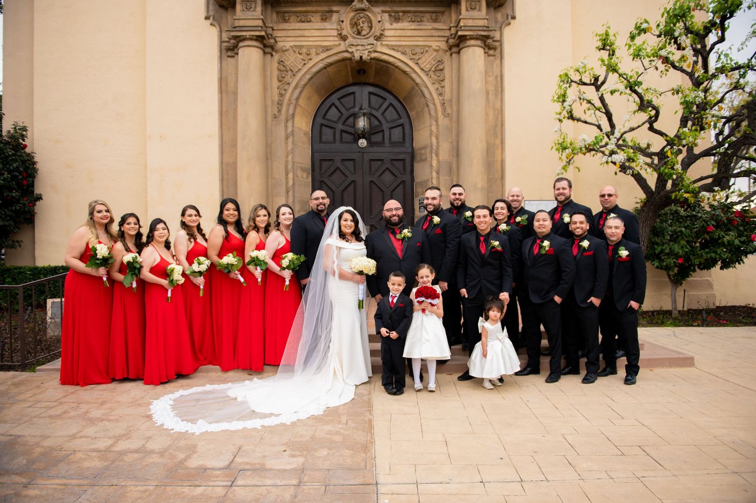 Wedding party in formal attire poses for group photos outside a grand stone church building.