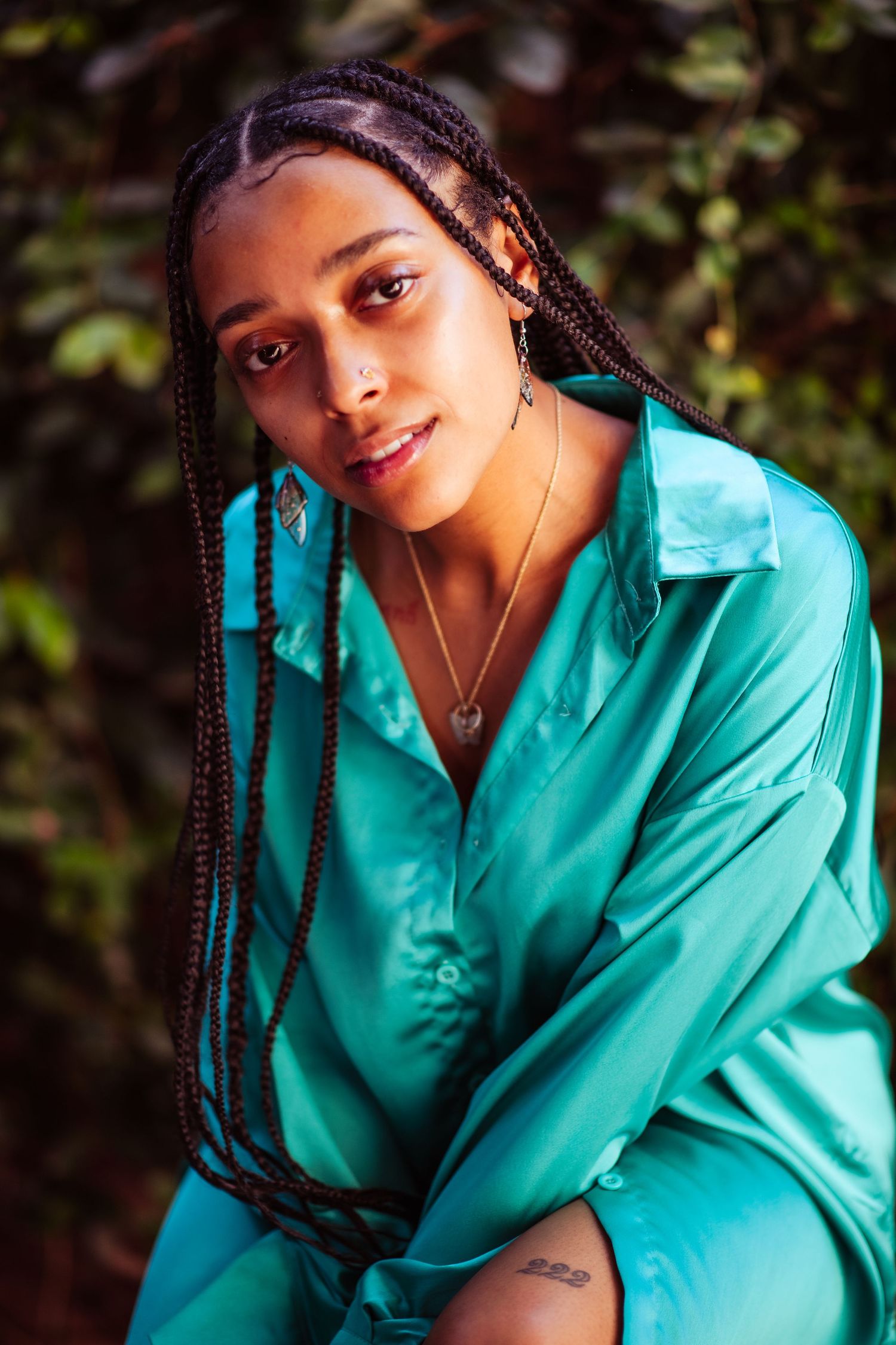 Person in turquoise satin shirt with long braided hair poses against greenery in warm sunlight.