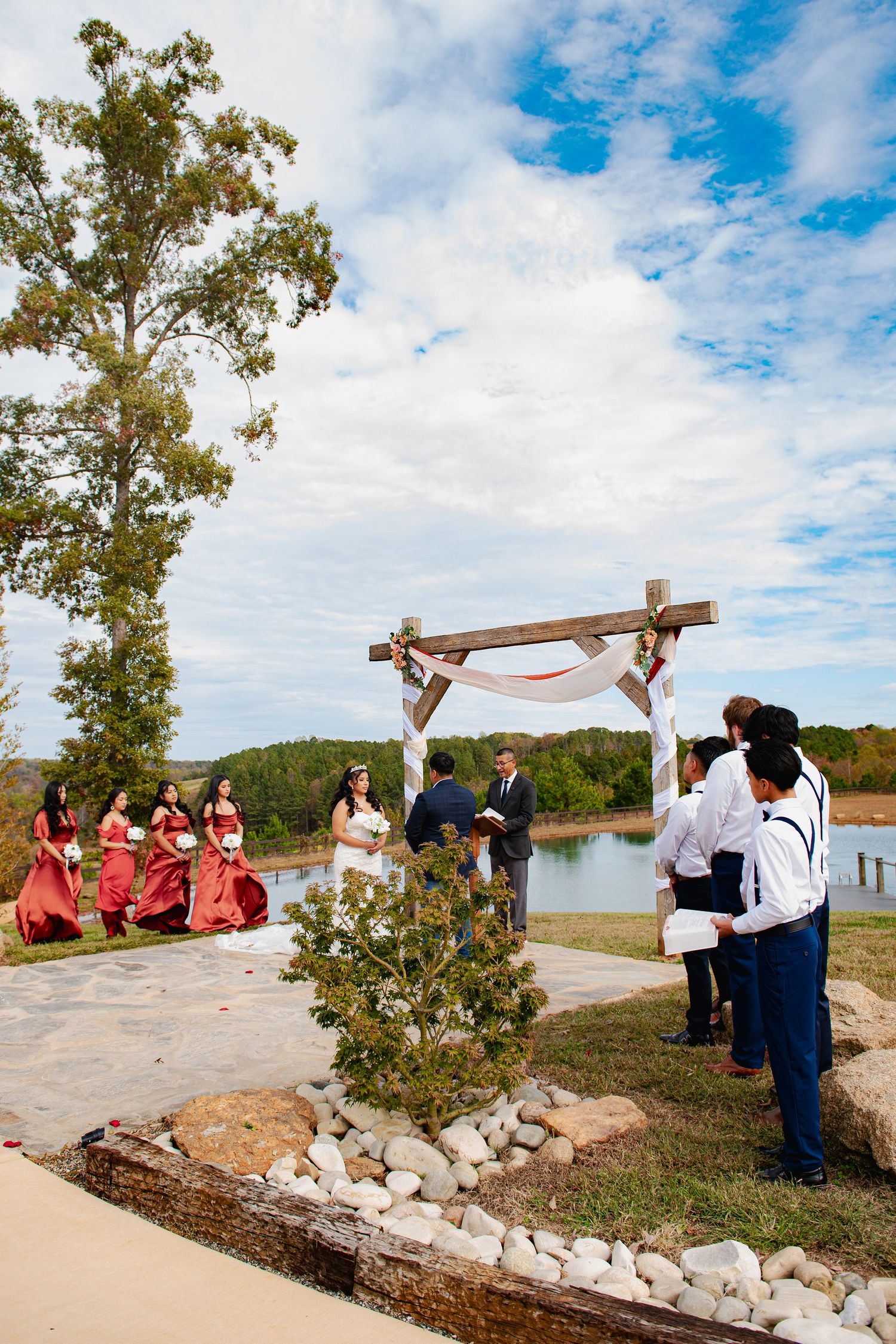 Outdoor wedding ceremony setup by lakeside with wooden arch and stone pathway under blue cloudy sky.