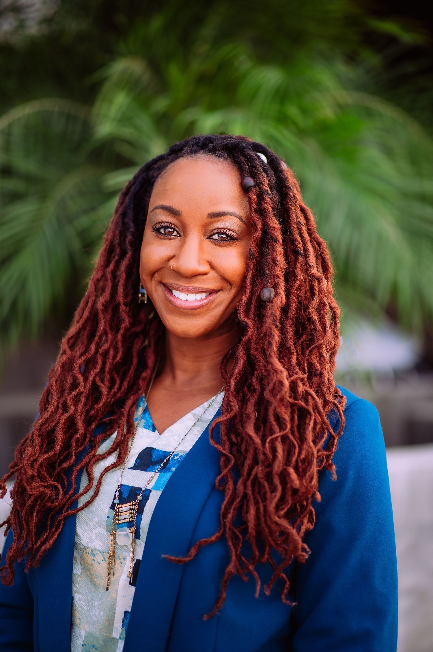 Professional headshots showing vibrant red locs hairstyle and bright blue blazer against natural outdoor background.