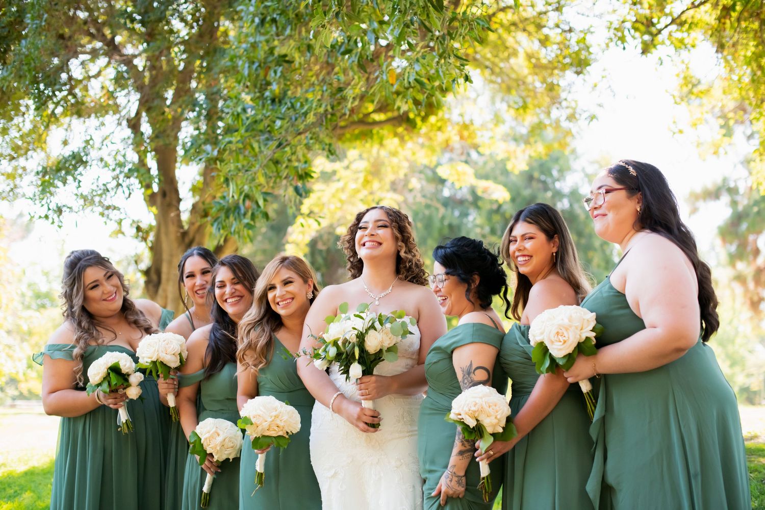 Wedding party poses in line wearing sage green dresses against sunlit outdoor backdrop.