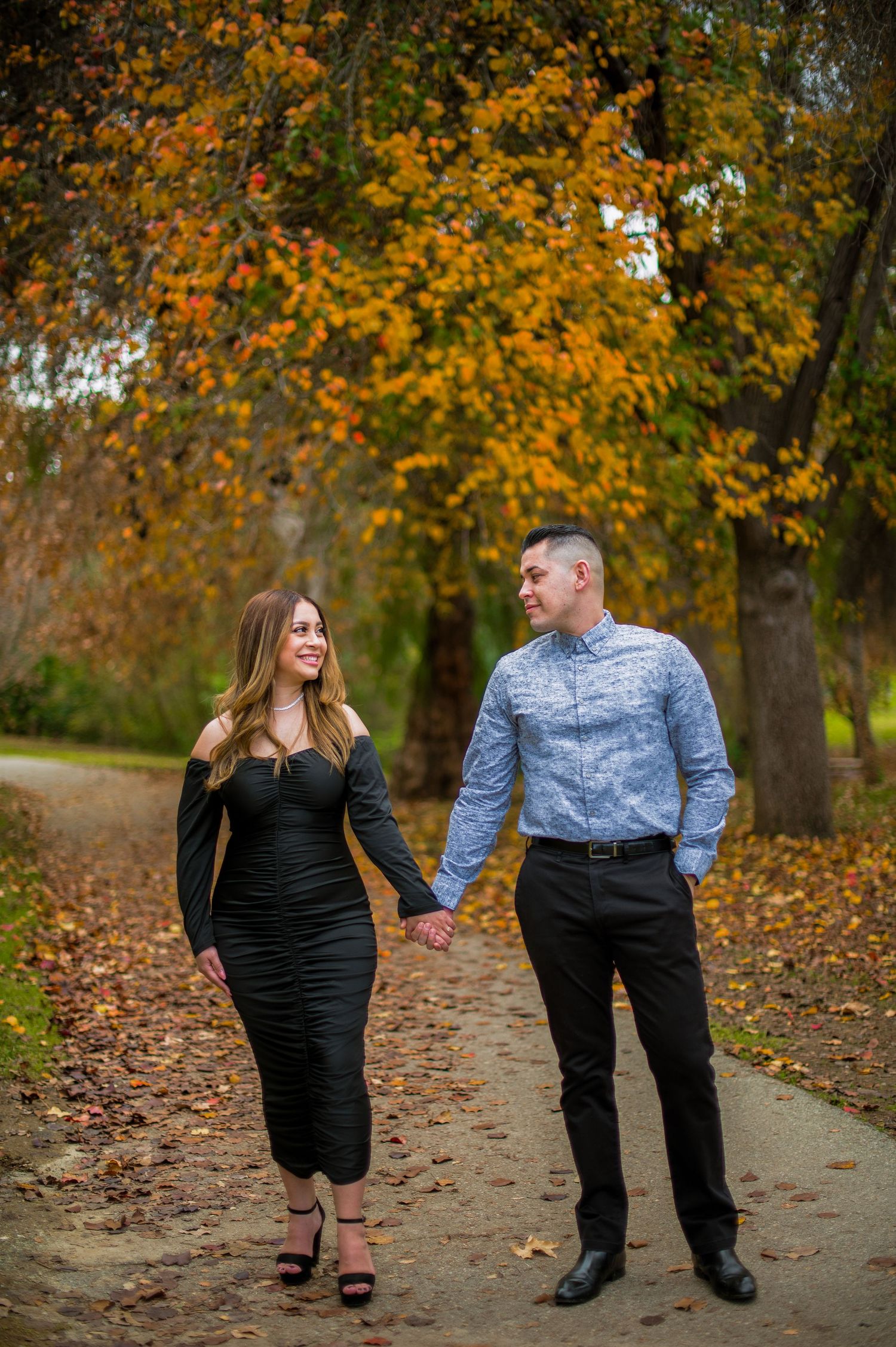 A couple shares a romantic autumn moment walking together on a leaf-covered path in a park.