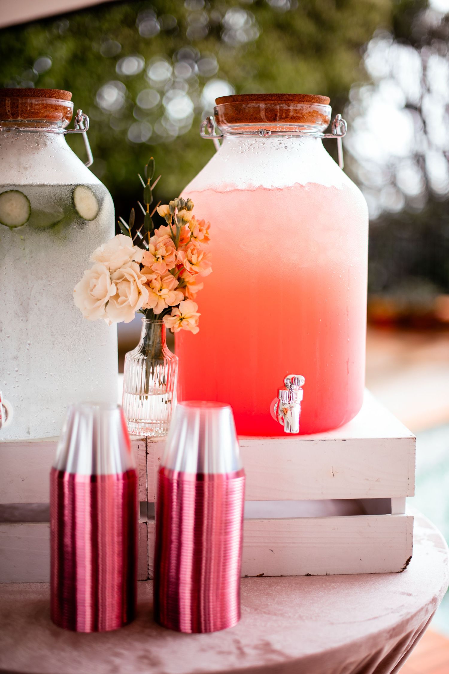 Large glass dispensers filled with pink beverages decorated with fresh flowers on a white table.