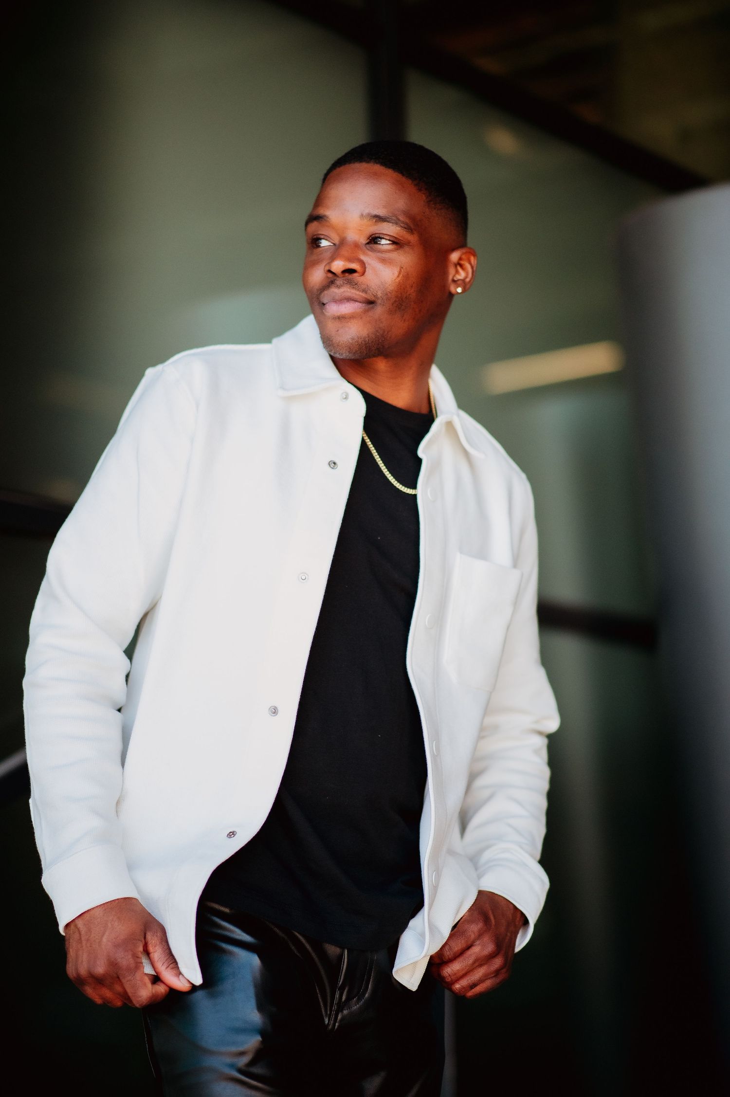 A person wearing a white jacket over a black shirt and blue jeans poses confidently in a casual indoor setting.