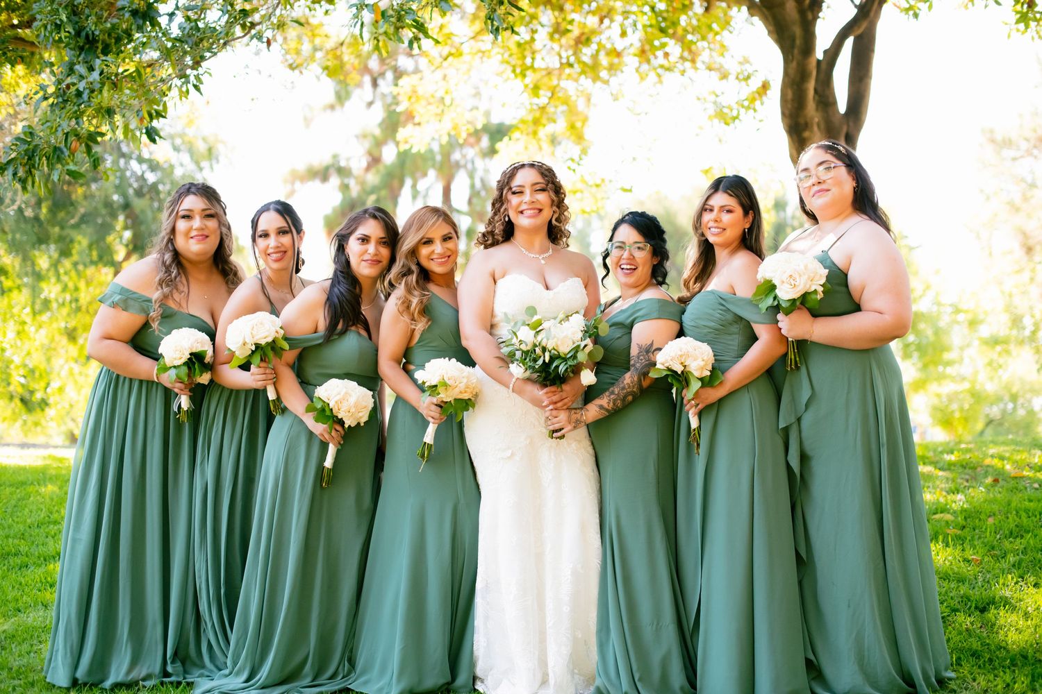 Bridal party in coordinating sage green dresses poses together on sunny grass lawn.